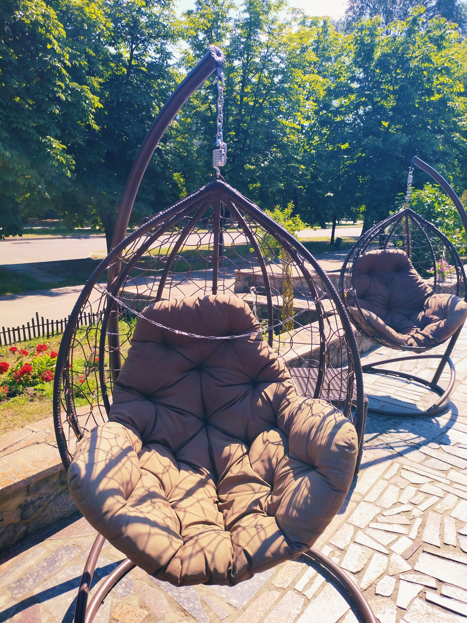 This image depicts an outdoor seating area in the city of Horishni Plavni, Ukraine, featuring two hanging basket chairs with brown cushions. The chairs are made of dark, woven material and suspended by metal stands and chains. The foreground chair is closer to the camera, showing its plush seat and the patterned shadow of tree branches cast upon it. The background chair is partially obscured by the foreground one, but its structure and cushion are still visible.
The chairs are placed on a paved ground made of irregularly shaped light-colored stones. To the left of the chairs, there is a low wooden fence bordering a patch of green grass and red flowers, suggesting a garden or landscaped area. Beyond the fence, a paved road and a sidewalk are visible, with trees lining the opposite side of the road. The background is dominated by lush green trees, indicating a sunny day with ample foliage. The lighting suggests it is daytime, with the sun casting distinct shadows. No people are present in the image.