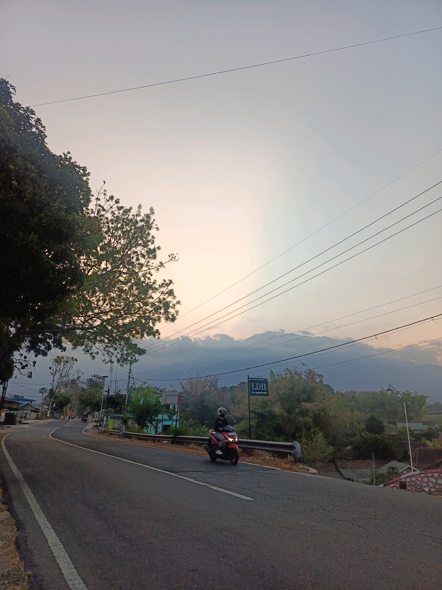 The image depicts a winding road in Campursari, Indonesia, under a hazy sky at dusk. A single red motorcycle with a rider wearing a helmet is traveling on the road. In the background, a sign with "LDII" and an arrow is visible. Trees and vegetation line the roadside, with some houses and buildings also present in the distance. Mountains are faintly visible in the background, partially obscured by clouds. Power lines are strung across the sky.