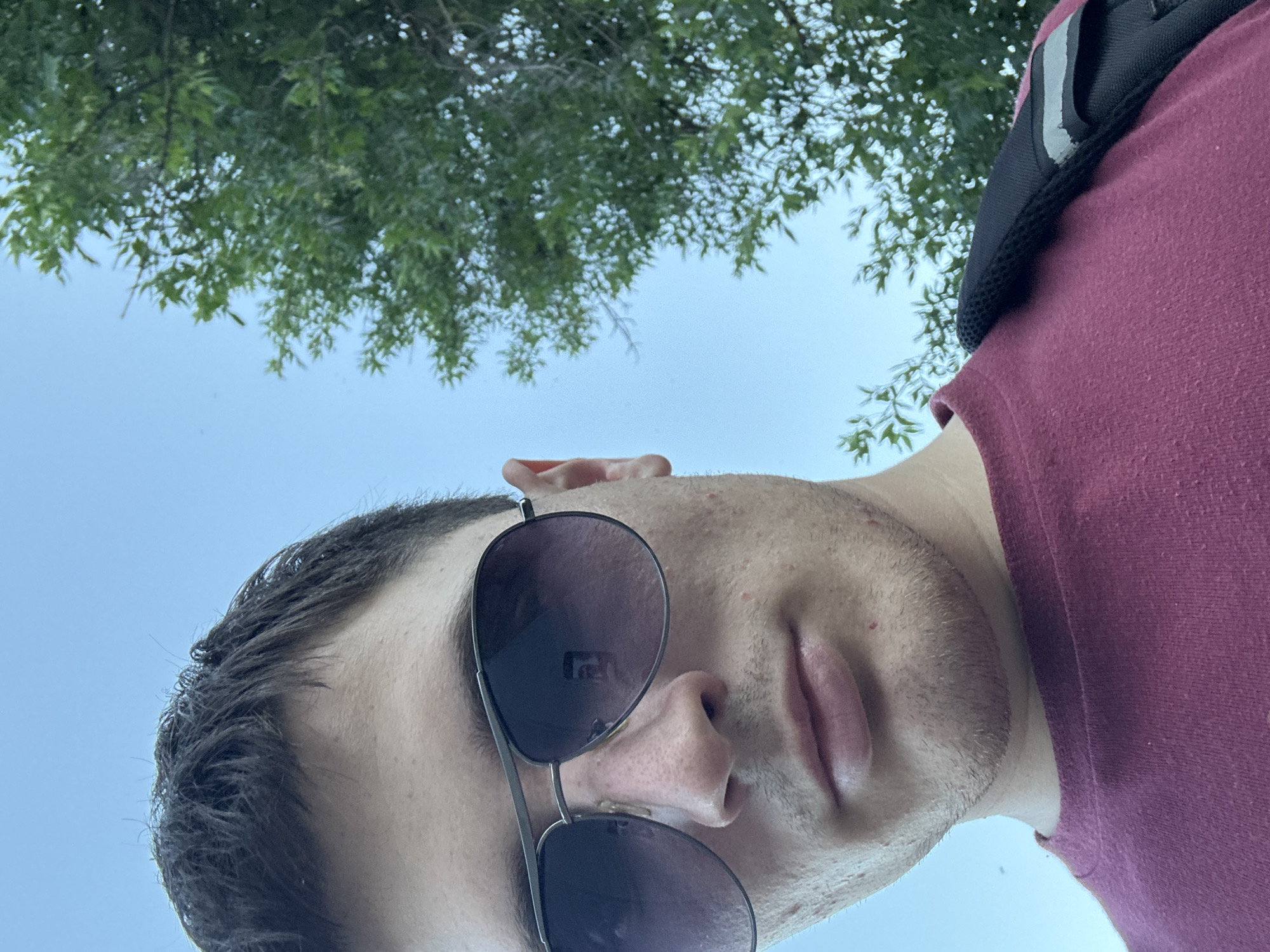 The image shows a close-up, low-angle, outdoor shot of a young man's face, partially obscured by aviator sunglasses. He is wearing a maroon t-shirt and a dark backpack strap is visible on his right shoulder. In the background, there is a clear blue sky and the green foliage of a tree. The scene is not identifiable as being in Stara Zagora, Bulgaria, or any specific city. The overall lighting suggests it is daytime.