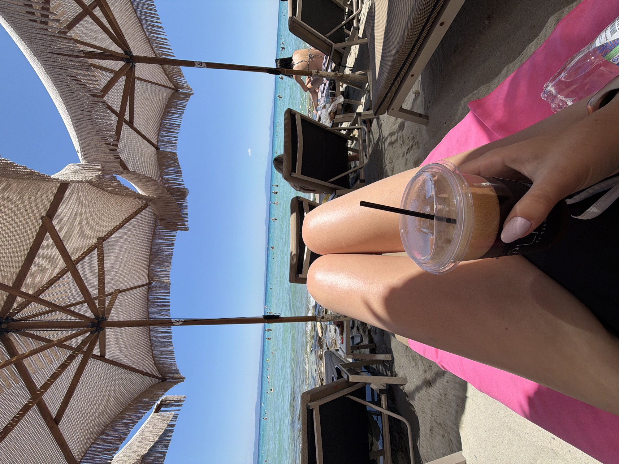 A first-person perspective shot on a sunny day at a beach in Kassandra Municipal Unit, Greece. The foreground features a person's legs extended on a pink beach towel, holding a to-go cup of coffee. In the middle ground, two beach chairs are visible, one with a person seated in it. The background shows the turquoise sea with several people swimming and enjoying the water, and the horizon is a light blue sky. Several umbrellas with fringe detailing are set up to provide shade.