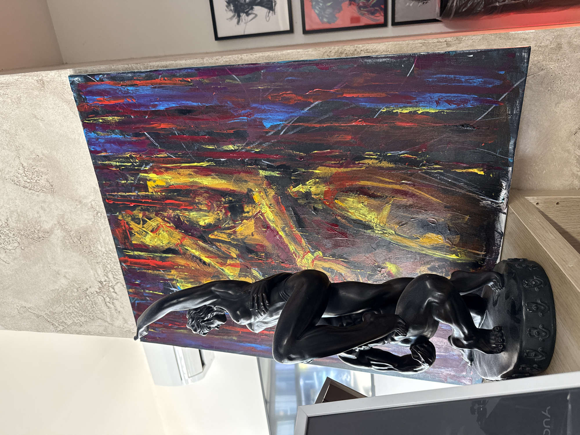 A medium shot depicts an abstract painting behind a black sculpture of two men wrestling, likely indoors. The painting features broad strokes of color, predominantly blues, purples, yellows, and reds, creating a textured and dynamic visual field. In front of the painting is the sculpture, which appears to be made of black metal or a similar dark material. The sculpture's details show two figures in a grappling position. The environment suggests a residential or commercial space, with elements of art and décor. The specific location in Sofia, Bulgaria, cannot be determined from the visual information alone.