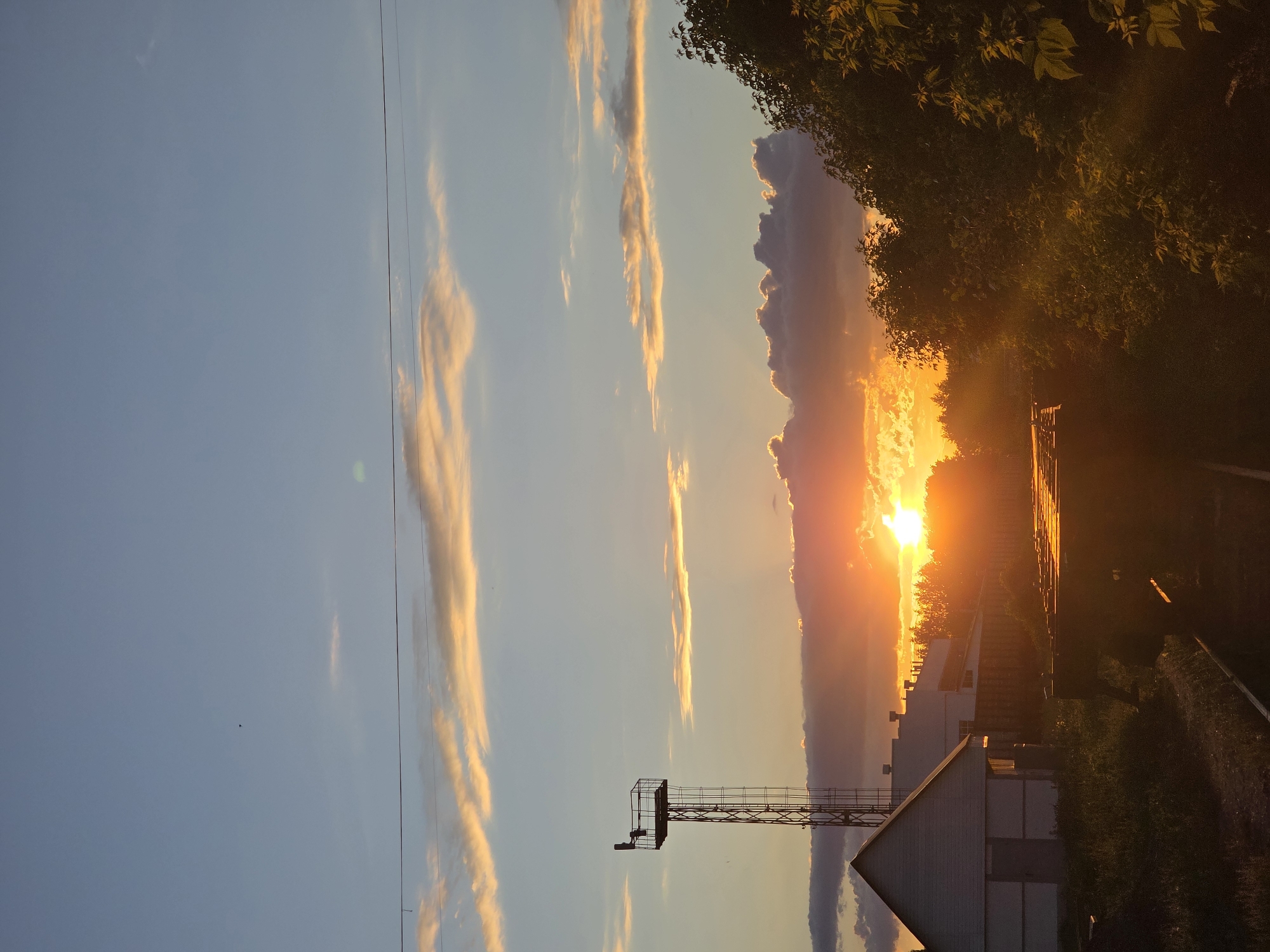 This image captures a serene sunset over a landscape in Chaikovsky, Russia. The sun, a brilliant orange and yellow orb, is prominently positioned low on the horizon, partially obscured by a band of dark clouds and distant foliage, casting a warm glow across the scene.
The sky transitions from a soft blue in the upper left to vibrant oranges and yellows near the horizon, illuminated by the setting sun. Scattered across the sky are elongated clouds, some appearing as bright, golden streaks reflecting the sunlight, while others are darker, silhouetted against the brighter parts of the sky. Thin power lines or wires are faintly visible stretching across the upper left quadrant.
In the foreground and mid-ground, various elements are silhouetted against the bright sky. Dense, dark green foliage, likely trees, frames the upper right and right side of the image, with some leaves subtly catching the golden light. Below the horizon, several buildings are visible. A prominent white building with a pitched roof is located in the lower left portion of the frame. Further to the right, other structures and what appears to be overgrown ground or bushes are present. A tall, metal lattice tower with a platform at its top stands out in the lower left, its structure sharply defined against the colorful sky.
There are no people or specific activities taking place. The overall impression is one of a peaceful end to the day, with the dramatic colors of the sunset being the central focus. No visible text is present in the image.