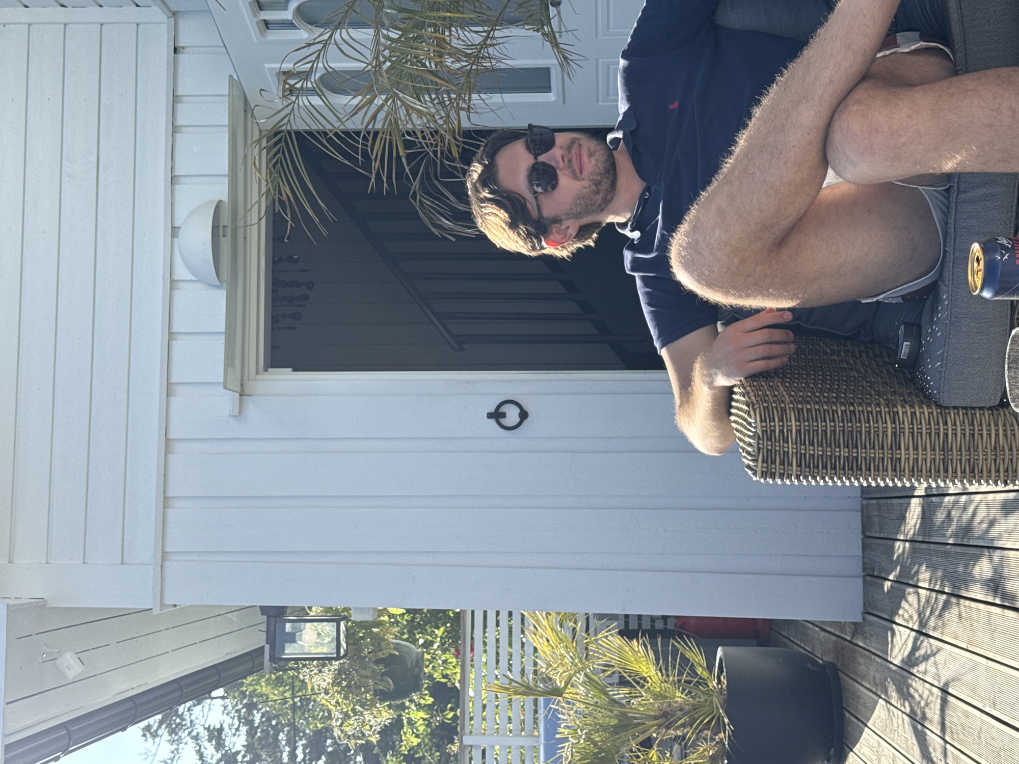 A young man with brown hair and sunglasses is sitting in a wicker chair on a wooden deck in front of a white wooden house. He is wearing a navy blue polo shirt and light-colored shorts. His legs are crossed, and his arms are resting on the arms of the chair. In the background, there is a doorway with a dark-colored door and stairs visible inside. There are also some potted plants and a fence. The scene appears to be on a sunny day. The location is Larkollen, Norway.