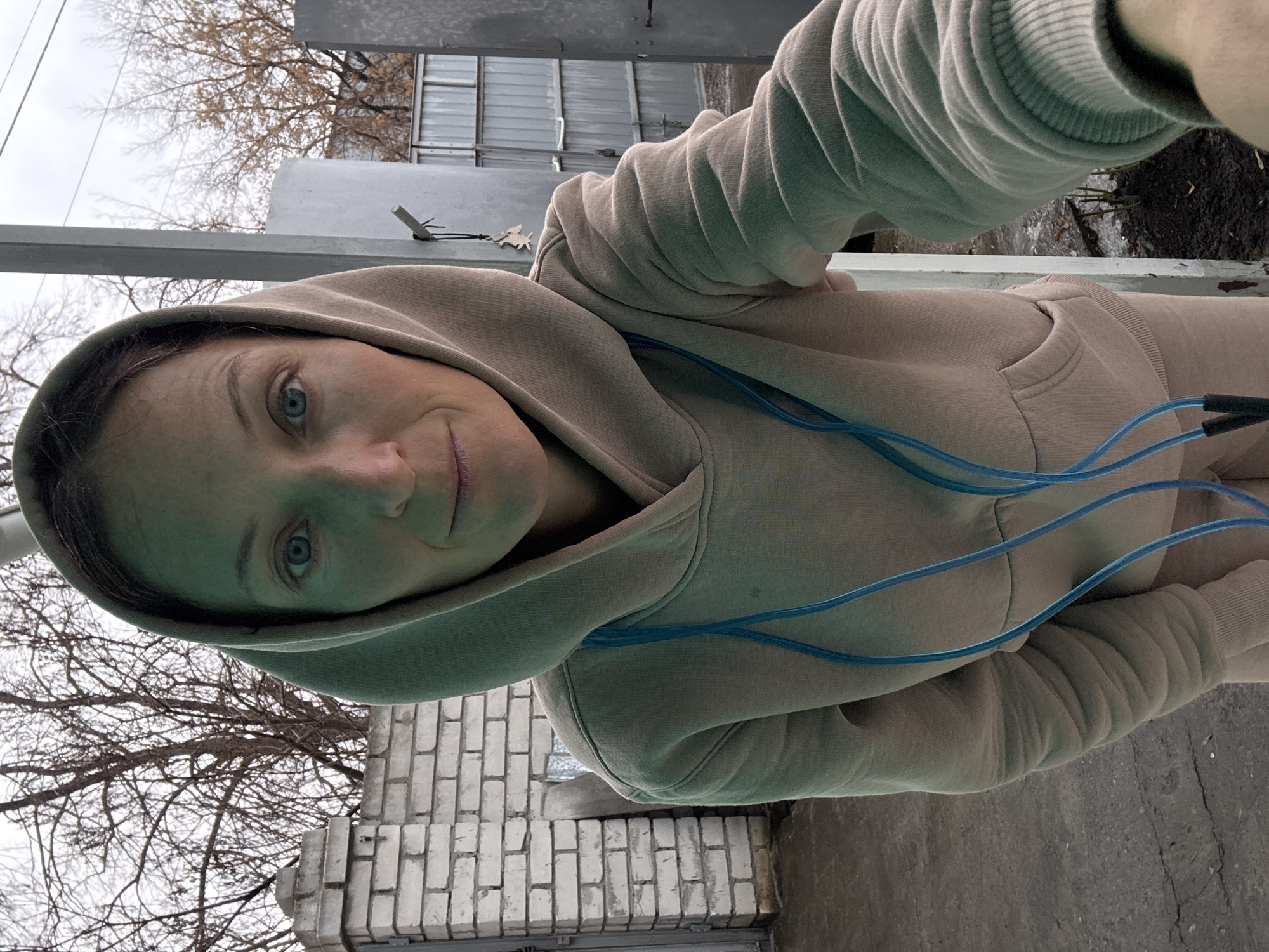A woman with blue eyes and a tan hoodie is taking a selfie. The location is in Ulyanovsk, Russia, as evidenced by the typical architecture and the general urban appearance. In the background is a metal fence and bare trees, indicating it might be late autumn or early spring. A brick wall and concrete ground are also visible.