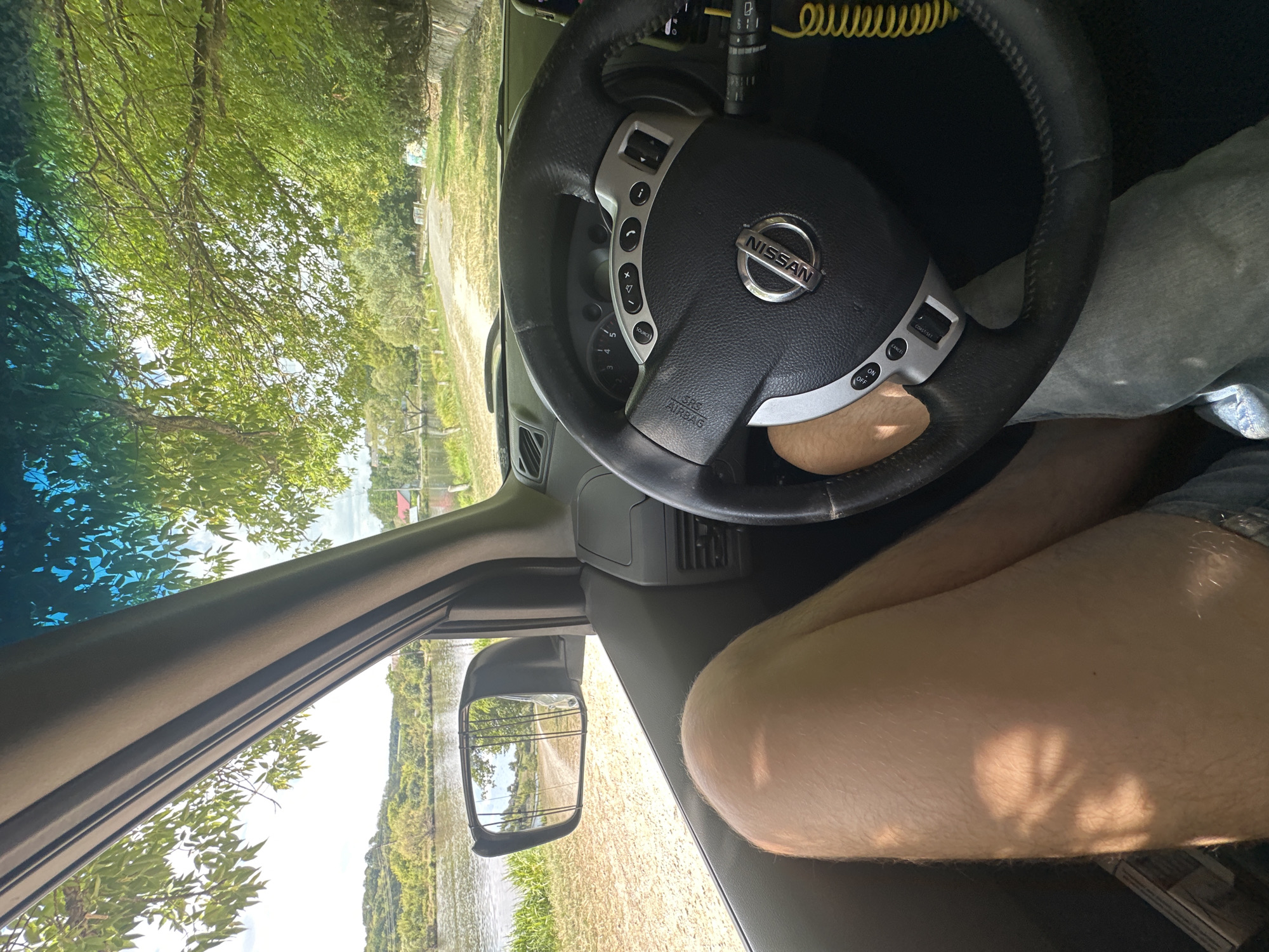 The image shows the interior of a Nissan vehicle, looking out through the driver's side window and windshield. In the foreground, a person's bare legs are visible, with sunlight casting a shadow pattern on the right thigh. The steering wheel, with the Nissan logo, is centered in the frame. Outside the vehicle, a dirt road runs alongside a body of water, possibly a river or lake, with lush green trees and vegetation lining the banks and a distant hillside with some buildings. The scene appears to be in a rural or semi-rural area, likely Makiv, Ukraine, given the context. The sky is partly cloudy.