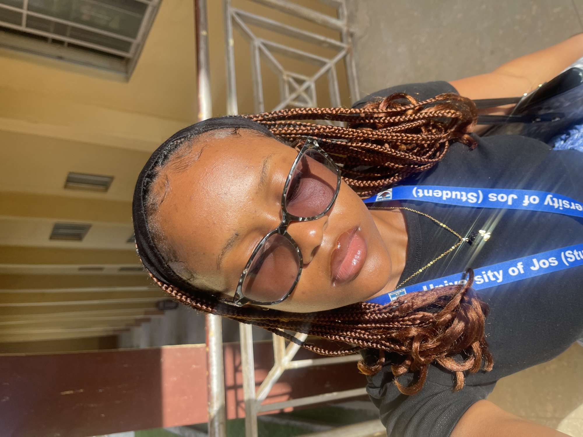A close-up, selfie-style portrait shows a young Black woman wearing a black t-shirt and sunglasses. Her hair is styled in individual braids with curly ends, and she has a black headband on. She is wearing a gold necklace with a small butterfly pendant. A blue lanyard, with the words "University of Jos (Student)" printed in white, hangs around her neck. She is standing in front of a building with yellow walls and what appears to be a covered walkway or balcony. The lighting suggests it is daytime, with sunlight casting shadows. The architectural style of the building with its repetitive windows and railings is consistent with institutional buildings often found in university campuses. Without specific identifying markers of the University of Jos campus, the exact location within Jos, Nigeria, cannot be determined from the image alone.