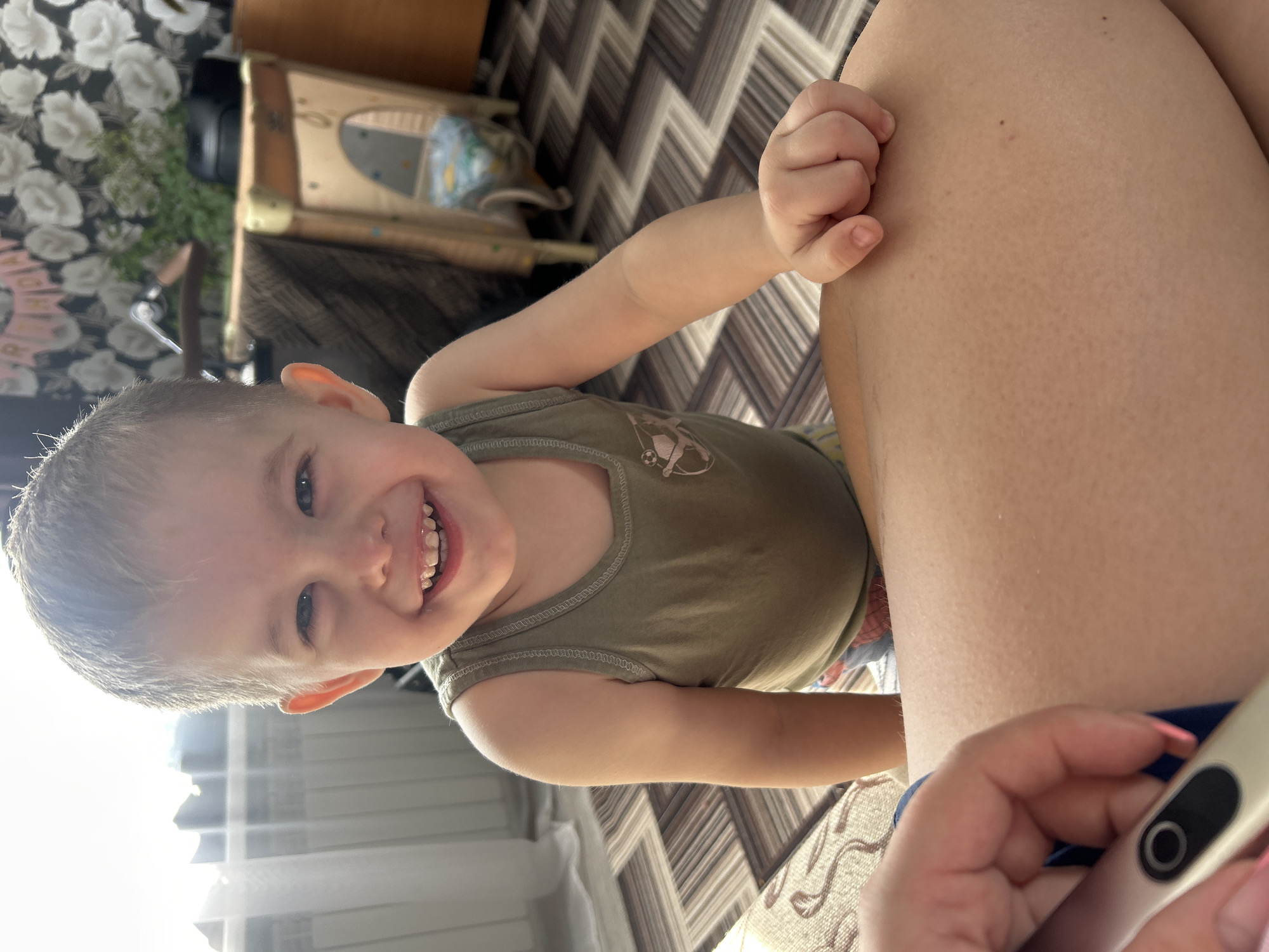 A child, approximately 2-3 years old, is smiling directly at the camera. He is wearing a green tank top. His left hand is near the leg of an adult. An adult is holding a thermometer. The background shows a patterned floor. The image was taken indoors, possibly in a residence in the city of Ольховка, Russia.