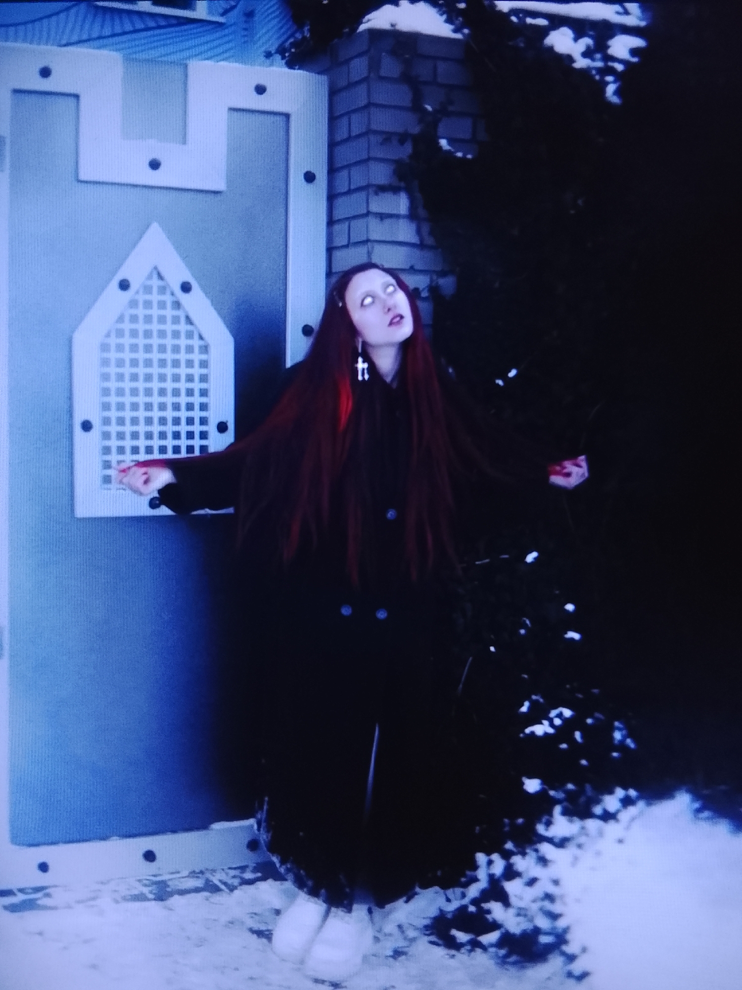 A young female with long, dark red hair and bright white eyes stands in front of a light blue, castle-like structure. She is wearing a long, dark coat and white fuzzy shoes. A cross pendant with two small black beads hangs from her neck. Her arms are outstretched, and her hands are held open. The area around her is covered in snow, and a dark, bushy tree with some snow on it is visible to her right. The background includes a brick wall and parts of a building. The overall scene has a dark and eerie atmosphere. The location is not specified as Prisel'e, Russia, or any other specific location.