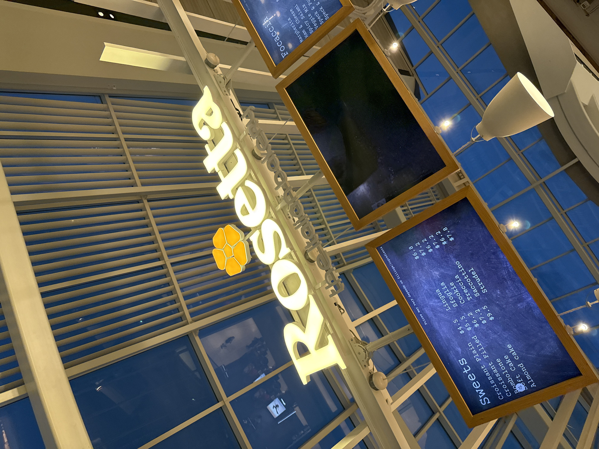 The image is taken indoors, showing the "Rosetta" signage and menu boards, likely a restaurant. The signage has a yellow flower-like symbol and the name is in glowing, white capital letters. Two menu boards are visible. One reads "Sweets" and lists items with prices such as "Croissant Plain $4.5", "Almond Cake $9.5", and "Strudel $7.8". The other menu board lists "Focaccia" items with names and prices. The background features a grid-like structure, presumably a window system that allows views of the sky. A white, cone-shaped light fixture is also visible. The angle of the shot is upwards, suggesting it was taken from a position looking up at the signage and menu boards. Given the presence of Italian menu items and the architecture, the image may have been taken in an airport terminal or an open shopping mall within the city of Miami, Florida, United States of America.