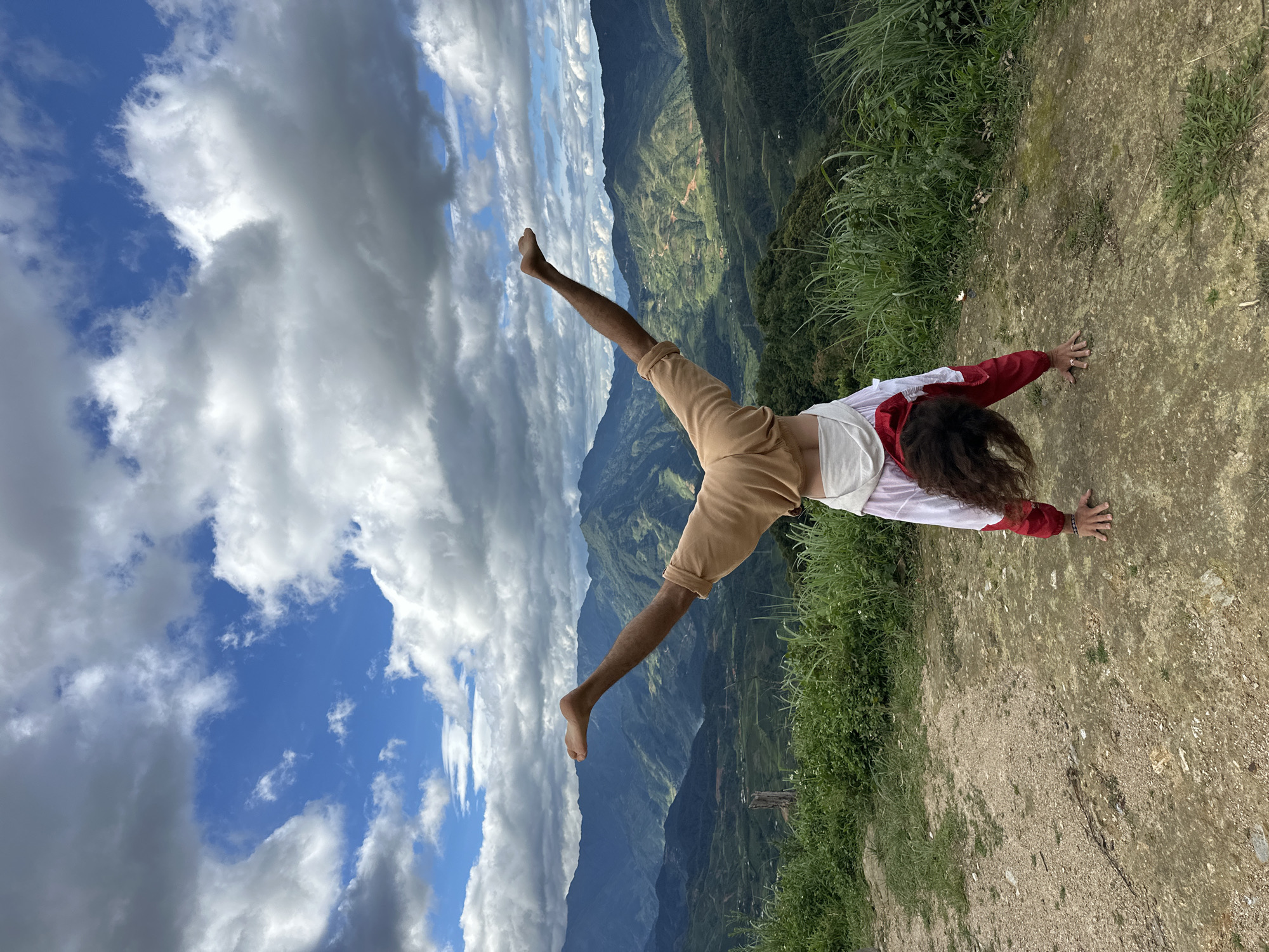 A single individual performs a handstand on a dirt and gravel path with hands placed on the ground. The person has long, dark, curly hair falling towards the ground and is wearing a red and white garment over what appears to be a light-colored top, and light brown or beige pants. Their bare feet are spread wide in an inverted V-shape against the sky. The immediate foreground consists of a mottled dirt and rocky surface with sparse green vegetation. Beyond the path, lush green foliage and grasses are visible. The midground and background feature a panoramic view of layered green mountains, some with visible terracing, under a partly cloudy sky with large white and grey clouds interspersed with patches of blue. The scene is located in Hanh Phuc, Vietnam.