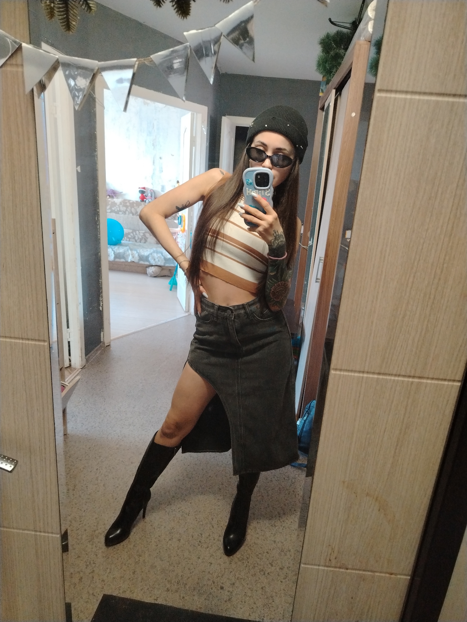 A full-length, indoor selfie features a slender woman wearing a black beanie, sunglasses, a striped crop top, a dark denim skirt with a high slit, and black heeled boots. She holds a cell phone with a light blue case up to her face, presumably taking the picture in a mirror. Her left arm displays a large tattoo. The room has a neutral color palette. Decorations, possibly party decorations, are suspended above a doorway. A bed can be seen in the room beyond. A wardrobe with wood-grain doors is visible in the background. The floor is a speckled light gray. The location is Noviy, Russia.
