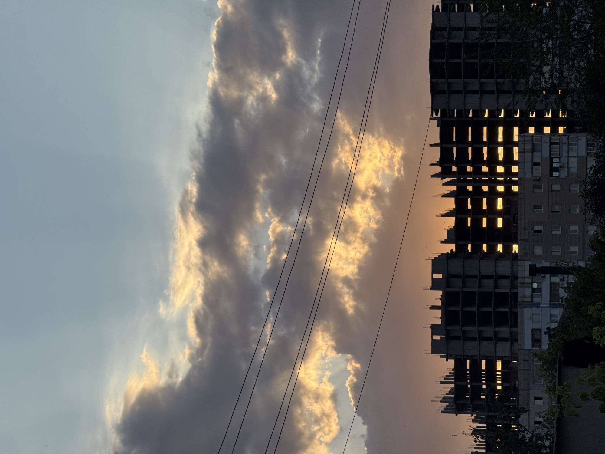 The image depicts an unfinished building against a sunset sky in Zaporizhzhia, Ukraine. The building, in a skeletal state with visible concrete structures and rebar, dominates the right side of the frame. Sunlight streams through the open floors and windows, creating bright rectangles of light against the dark, unfinished facade. To the left of the building, dramatic clouds are illuminated by the setting sun, casting golden and pink hues across the sky. Several power lines run diagonally across the frame, obscuring parts of the clouds and the building. The overall scene evokes a sense of ongoing construction and the beauty of a sunset. The time of day is clearly sunset, and the weather appears partly cloudy. There is no visible text in the image.