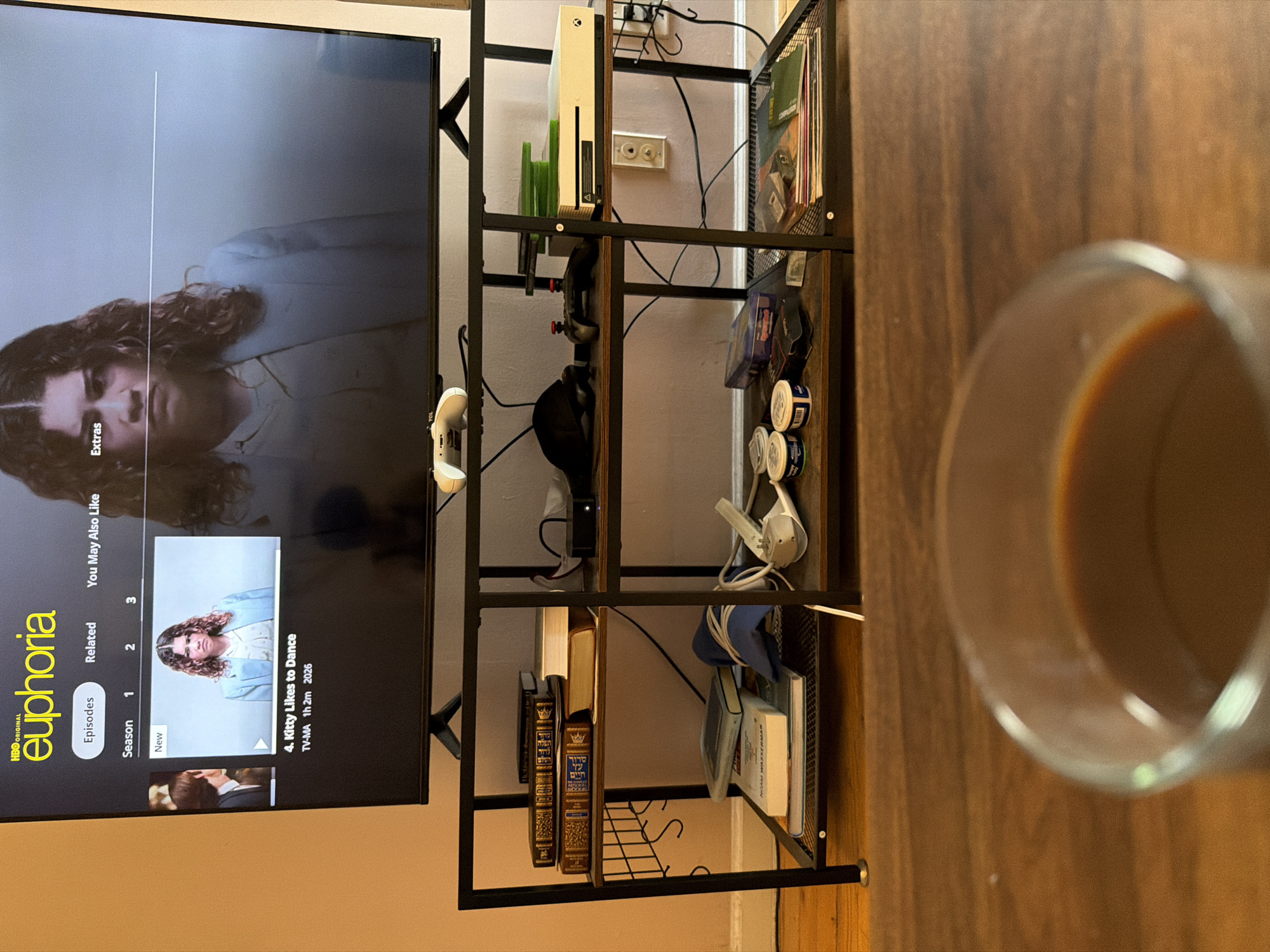 The image shows a living room interior. On the left, a television screen displays an episode of the HBO show "Euphoria," specifically "Kitty Likes to Dance." Adjacent to the television is a black metal shelving unit. This unit contains a white Xbox Series S console, game controllers, a headset, books (including what appears to be a religious text in Hebrew), and other miscellaneous items. To the right of the shelving unit, a partially visible glass of coffee sits on a wooden surface. The room is illuminated with indoor lighting.