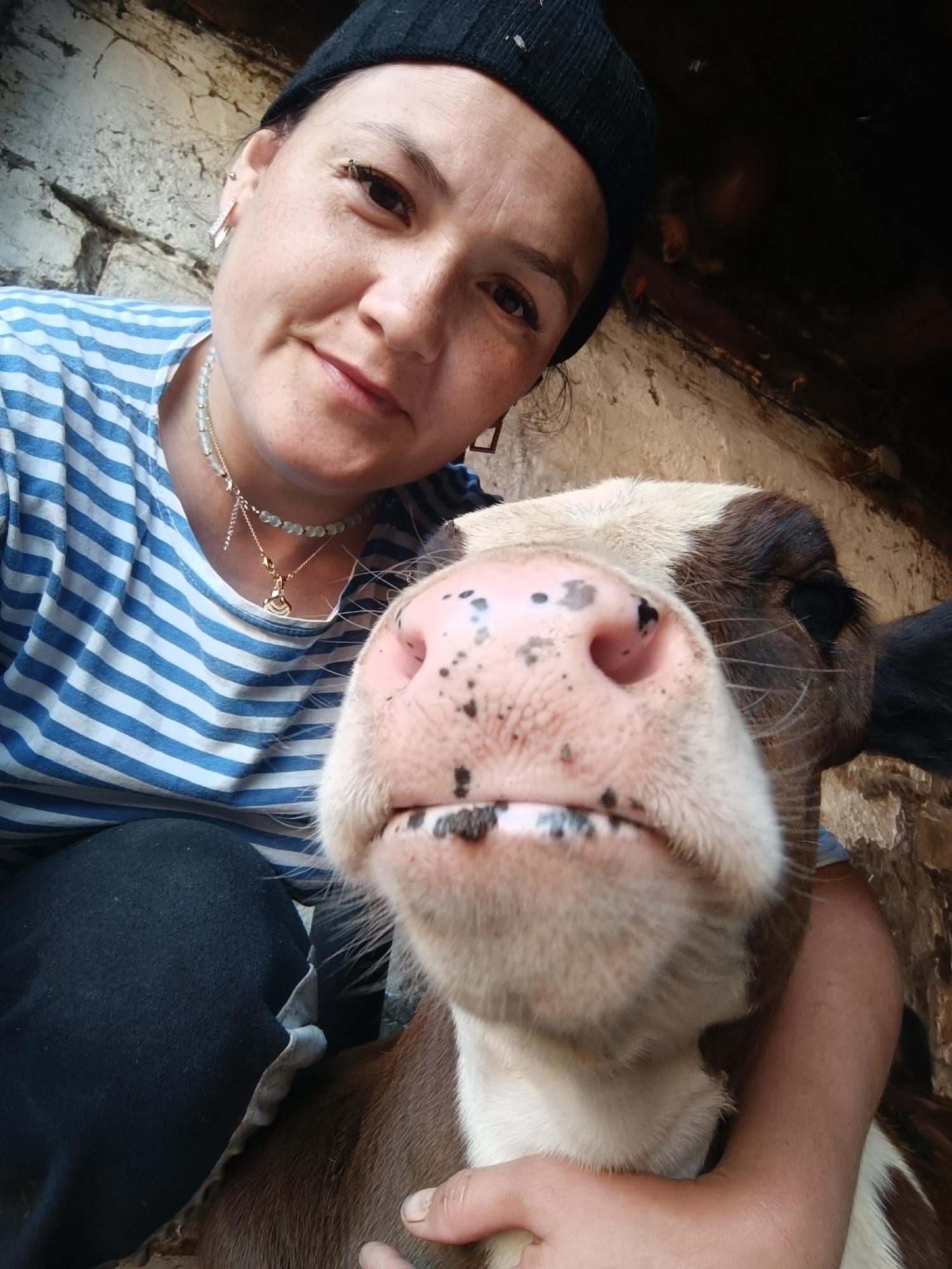 In a dimly lit, rustic environment, likely a barn or stable, a woman wearing a black beanie and a blue and white striped t-shirt is taking a selfie with a young calf. The woman's face is partially obscured by the calf's snout, which is positioned very close to the camera. She has light skin with freckles and is wearing earrings and necklaces. The calf has a white face with brown markings around its eyes and ears, and a pink nose with black spots. It appears to be a domestic setting, with rough stone and wooden textures visible in the background. The woman is holding the calf, suggesting an affectionate interaction. The lighting is natural and somewhat harsh, highlighting the textures of the subjects and the environment.