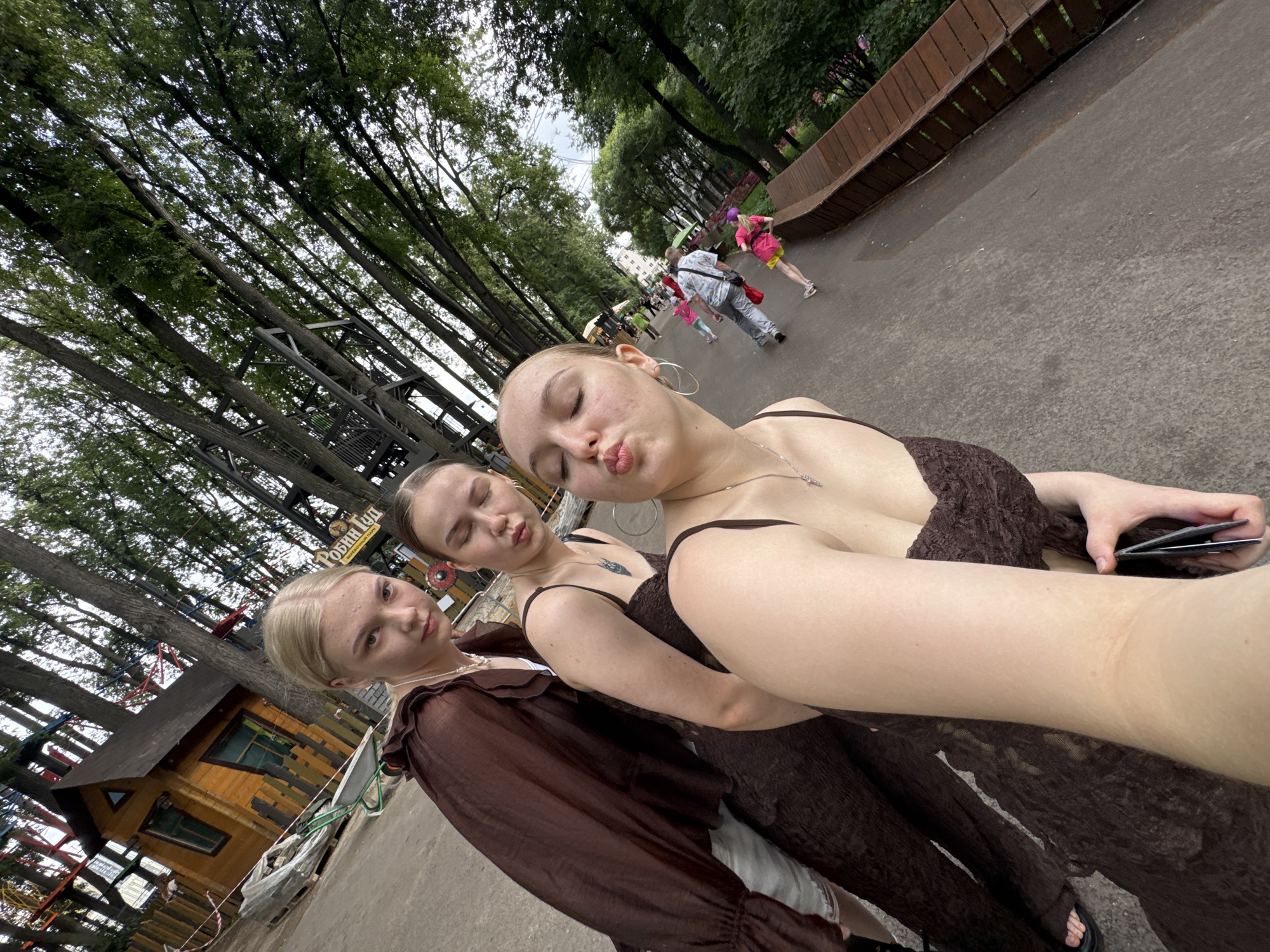 Three young women are taking a selfie outdoors in what appears to be an amusement park or recreational area. The woman closest to the camera, on the right, is making a kissing face with her eyes closed. She is wearing a dark brown dress and large hoop earrings. The woman in the middle, also with her eyes closed, is making a similar kissing face and is wearing a dark brown dress and a necklace. The woman on the left is looking at the camera with a neutral expression and is wearing a sheer brown top over a white garment, and appears to be wearing shorts.
In the background, there are tall trees, a wooden structure resembling a small building or attraction, and a path with other people walking. A sign with Cyrillic text is partially visible in the middle background. The overall environment suggests an outdoor public space with natural elements and man-made structures.
Based on the provided information, this location is likely in the city of Perm, Russia, possibly within a park or entertainment venue. The presence of Cyrillic text on signage would be consistent with this location.