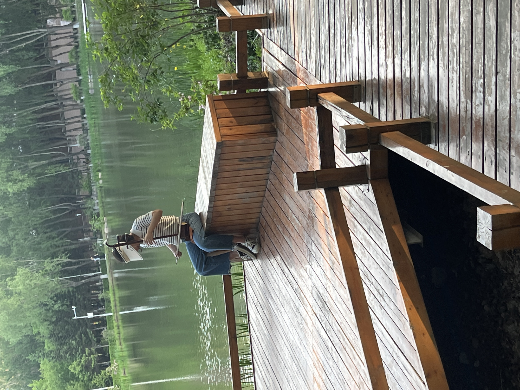 A man with a music stand is playing a two-stringed bowed instrument on a wooden deck next to a lake. The lake is surrounded by green trees and appears to be located in Changchun City, China. The man is wearing a striped shirt and jeans. He is facing the lake. The wooden deck has railings.