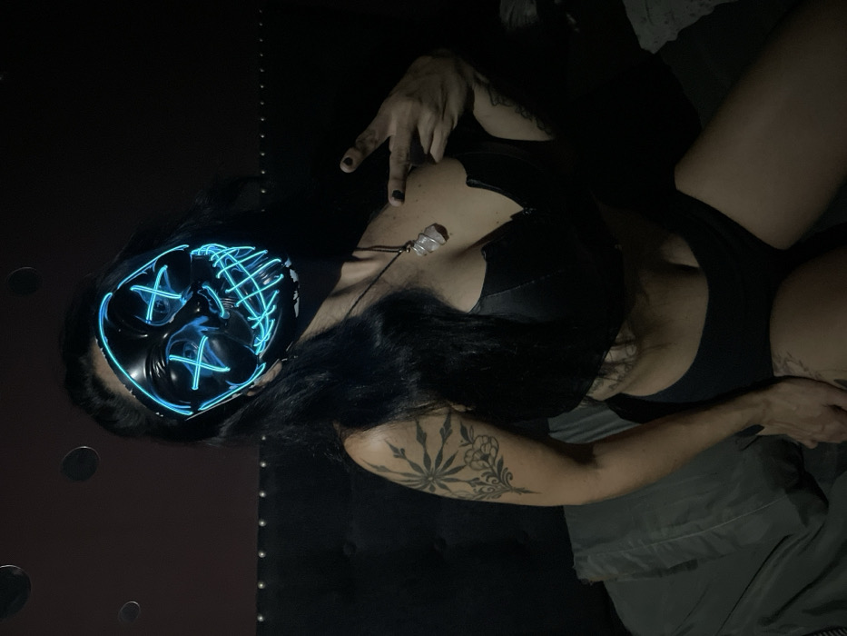 A person in a black bra and shorts lies on a bed, wearing a black mask with blue neon wiring. They have long, dark hair and tattoos on their arm and chest. A crystal pendant hangs around their neck. The background is dark, with a tufted headboard and scattered circular objects. Their hand is raised, fingers slightly curled.