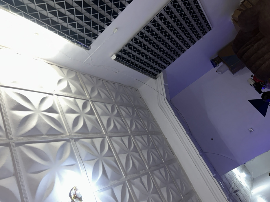 A modern interior showcases a textured white wall with repeating floral patterns, contrasted by geometric ceiling panels in black and white. A decorative wall molding accents the corner. The adjacent wall and ceiling are painted in a gradient of white to light purple. Part of a brown upholstered piece of furniture and some other objects are visible in the upper right corner of the image.