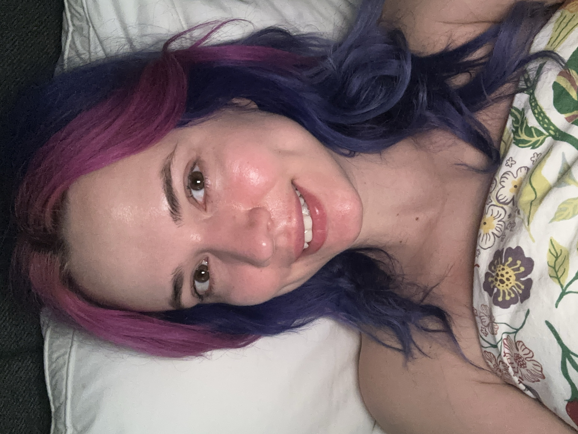 A fair-skinned woman with purple and blue hair lies on a pillow and floral-patterned sheets. She is looking at the camera with a slight smile. The location cannot be determined from the image.