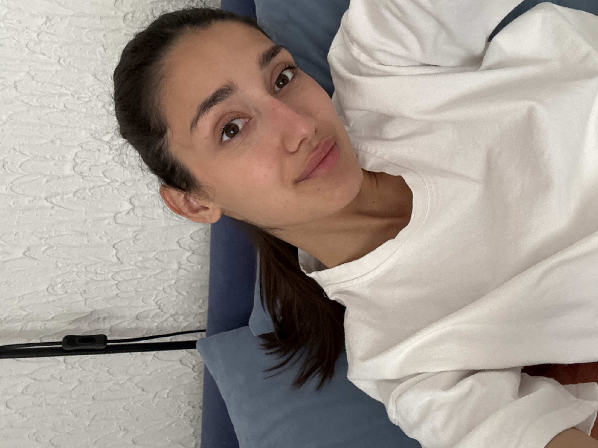 A woman with dark hair pulled back in a ponytail is lying on a blue pillow. She is wearing a white long-sleeved shirt. Behind her is a white textured wall with a black power cord running along it. The location is Sutomore, Montenegro.