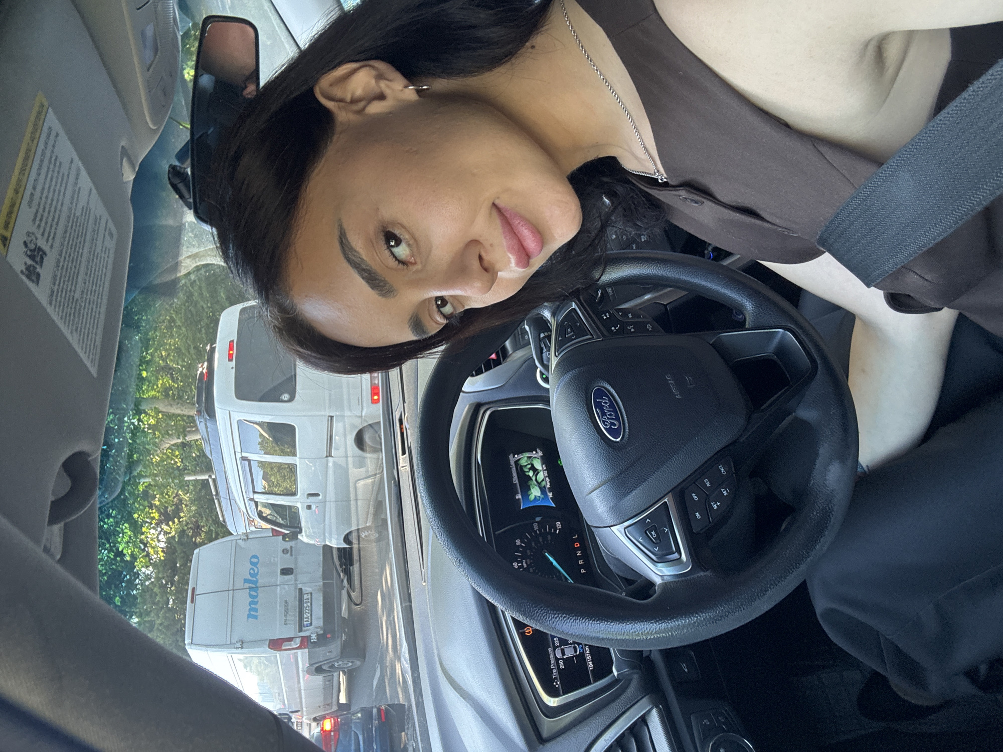 A woman sits in the driver's seat of a Ford vehicle in traffic on a sunny day in Tbilisi, Georgia. She is looking towards the camera with a slight smile. She has dark hair and is wearing a brown sleeveless top and a silver necklace. Her right hand is on the steering wheel, and her seatbelt is fastened. The car's dashboard is visible, showing the speedometer, tire pressure, and a digital display. Outside the car, other vehicles, including vans, are visible in traffic. Trees and foliage line the street. A warning label is attached to the car's visor. The image appears to be a selfie taken from inside the car.