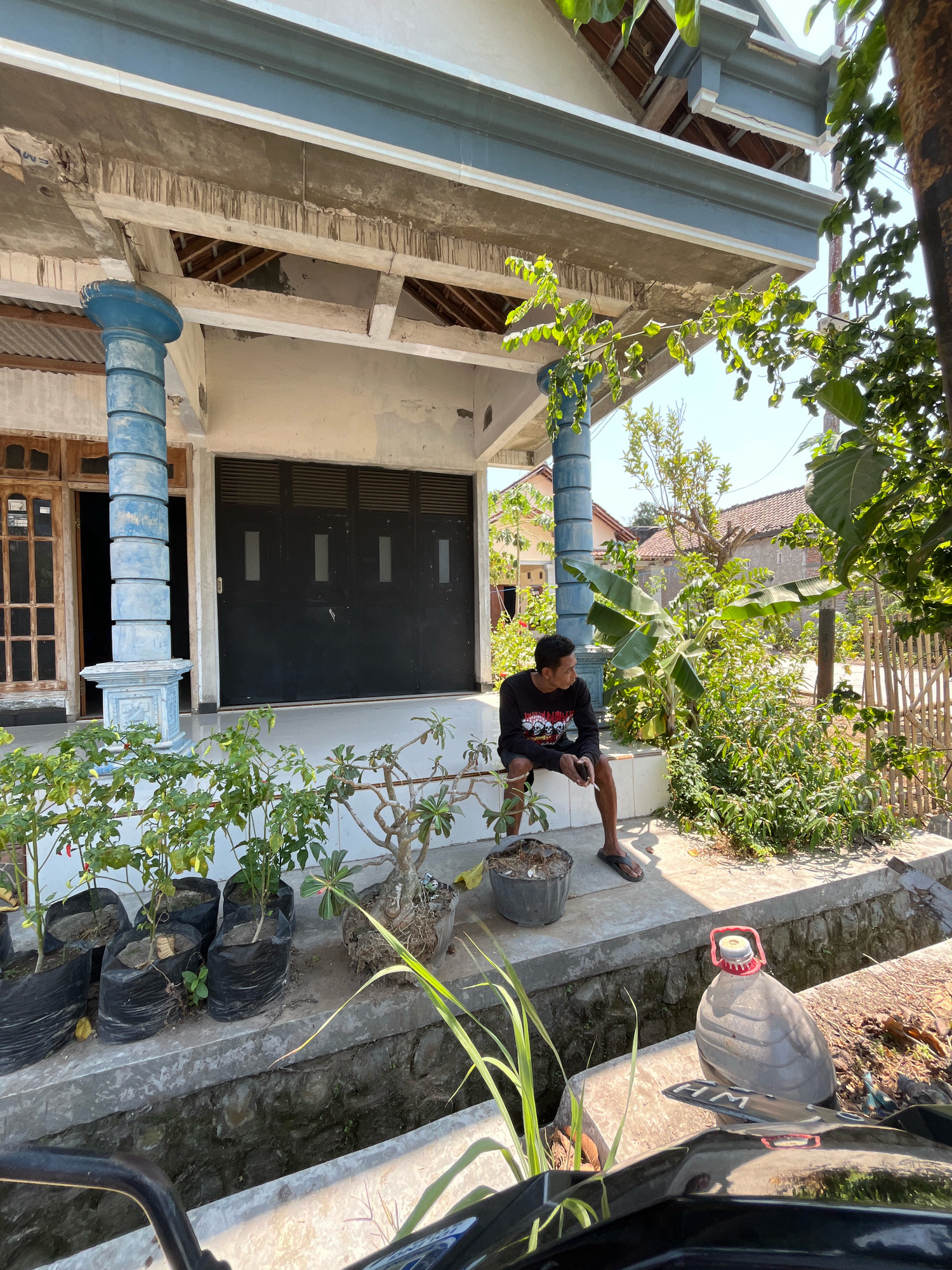 In Plosojenar, Indonesia, a young man sits on a concrete ledge outside a building with a dark, multi-paneled garage door and wooden-framed windows. He wears a long-sleeved black t-shirt with a graphic print and shorts, and flip-flops. He holds a cigarette in his right hand and a smartphone in his left. Several potted plants, including chili plants in black plastic bags, line the ledge. To the right of the man, a large plastic bottle with a red handle and cap sits on a cement channel. The scene is bathed in sunlight, with shadows cast by the building and surrounding vegetation, including banana trees and other leafy plants. The building's architecture features blue cylindrical pillars supporting an extended roof. In the background, residential structures with tiled roofs are visible.