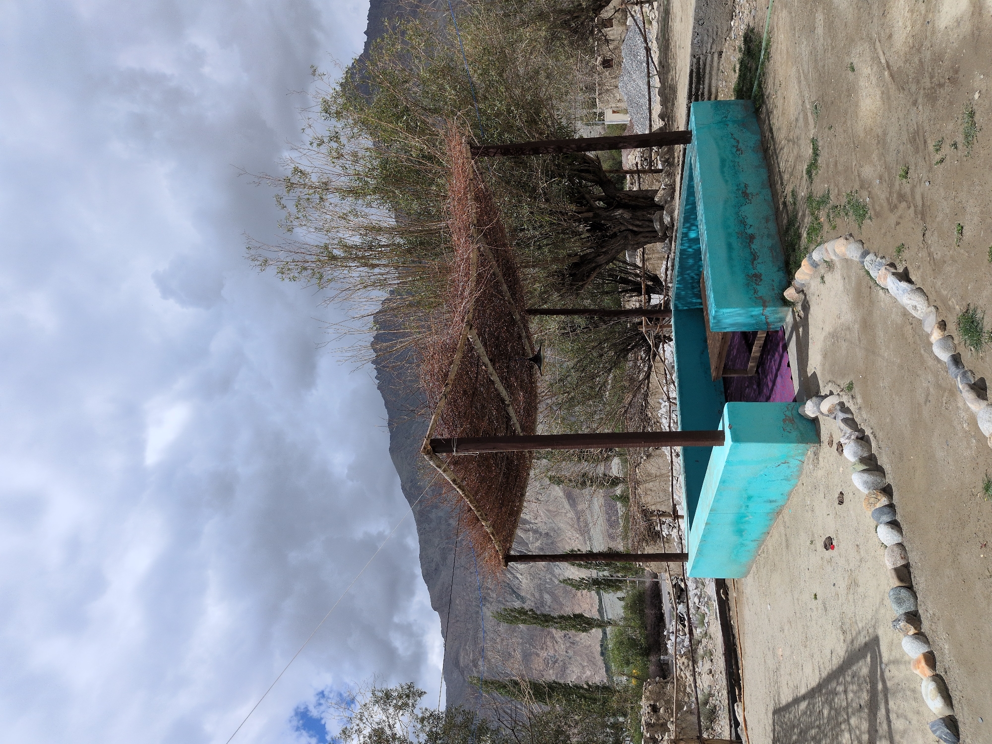 The image depicts an outdoor seating area in Panamik, India, characterized by a turquoise-colored, low-lying structure with a wooden bench inside, covered by a rustic pergola made of branches and twigs. The scene is set against a backdrop of arid, mountainous terrain with scattered trees and a cloudy sky. The ground is sandy and dusty, with a border of smooth, rounded stones lining a pathway. The overall impression is one of a simple, weathered structure in a remote, natural setting.