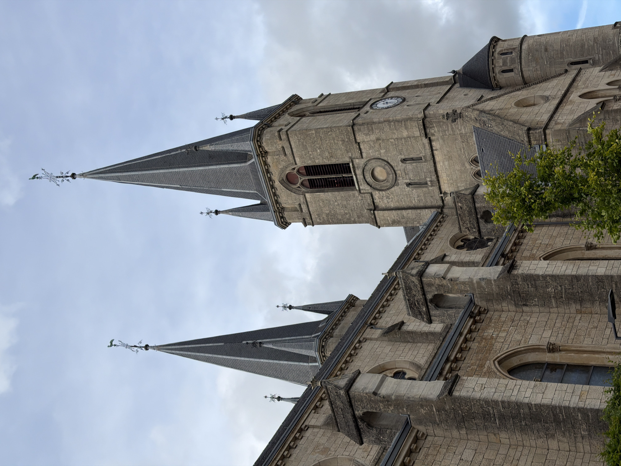 The image captures an upward, angled view of the St. Joseph's Church in Esch-sur-Alzette, Luxembourg. The architecture is predominantly Gothic Revival, featuring grey stone walls, pointed arches, and steeply pitched roofs. The church's steeple dominates the frame, topped with a weather vane. A clock face is visible on the tower. Adjacent to the church, a section with arched windows is visible, partially obscured by green foliage. The sky is overcast. The perspective is from below, emphasizing the height and grandeur of the structure.