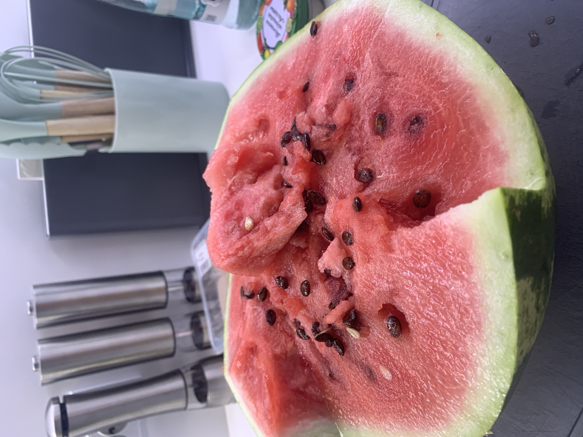 A close-up, low-angle shot shows a large slice of watermelon with visible black seeds and some of its pink flesh removed. In the background, out of focus, are silver salt and pepper grinders, a light blue cup holding kitchen utensils with wooden handles, and a clear bottle of liquid. The scene is indoors, likely in a kitchen setting. The image is undated and unsigned. The location is specified as Kolpino, Russia.