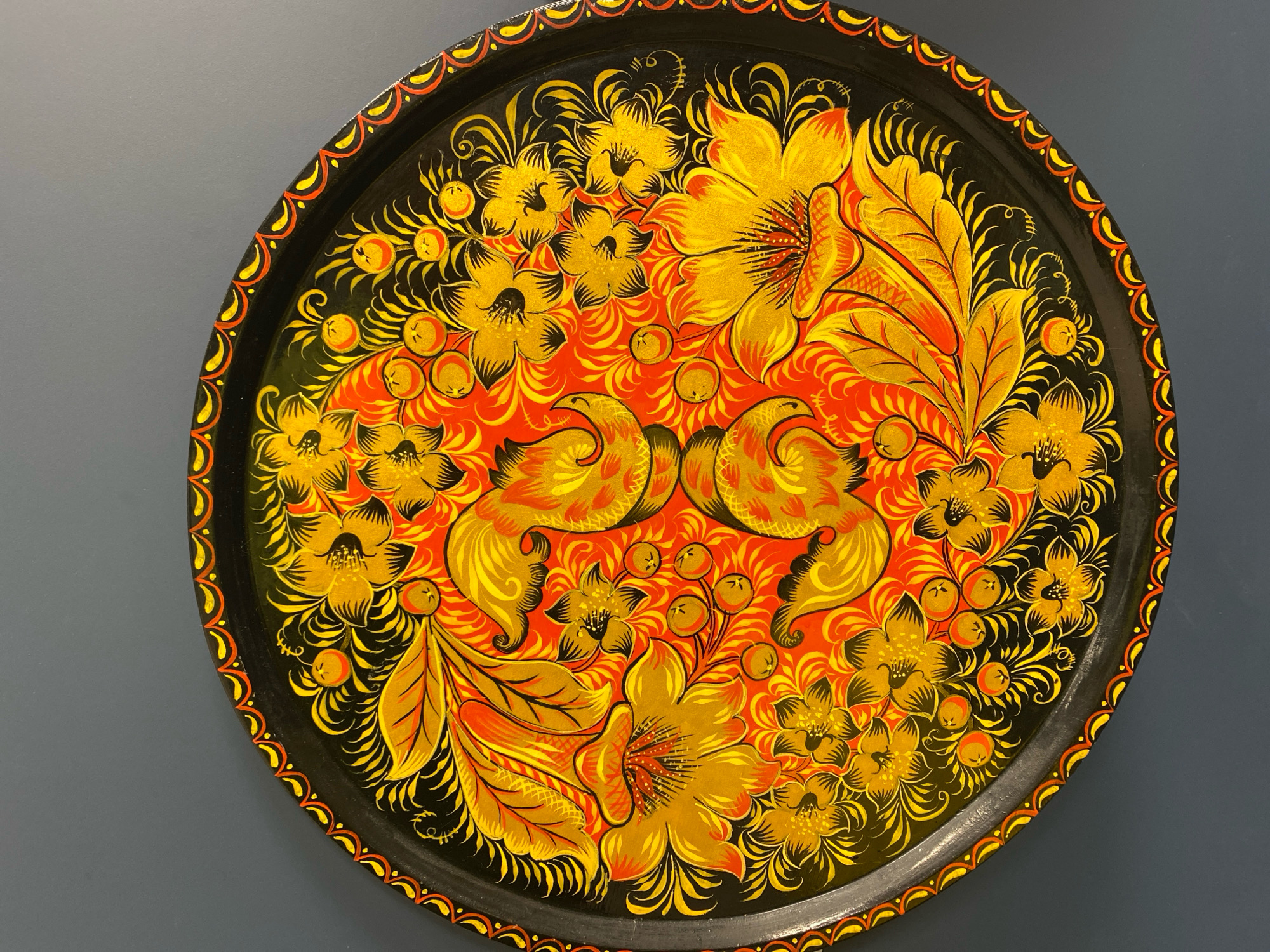 The image shows a circular tray with a traditional Khokhloma painting on a dark background. The painting features a central motif of two golden birds facing each other, surrounded by an intricate pattern of golden flowers, leaves, and red berries. The border of the tray is black with a decorative red and yellow pattern. The tray is set against a plain, dark gray wall. The location of this image is not specified in the provided context, and therefore cannot be linked to Ivanteyevka, Russia.