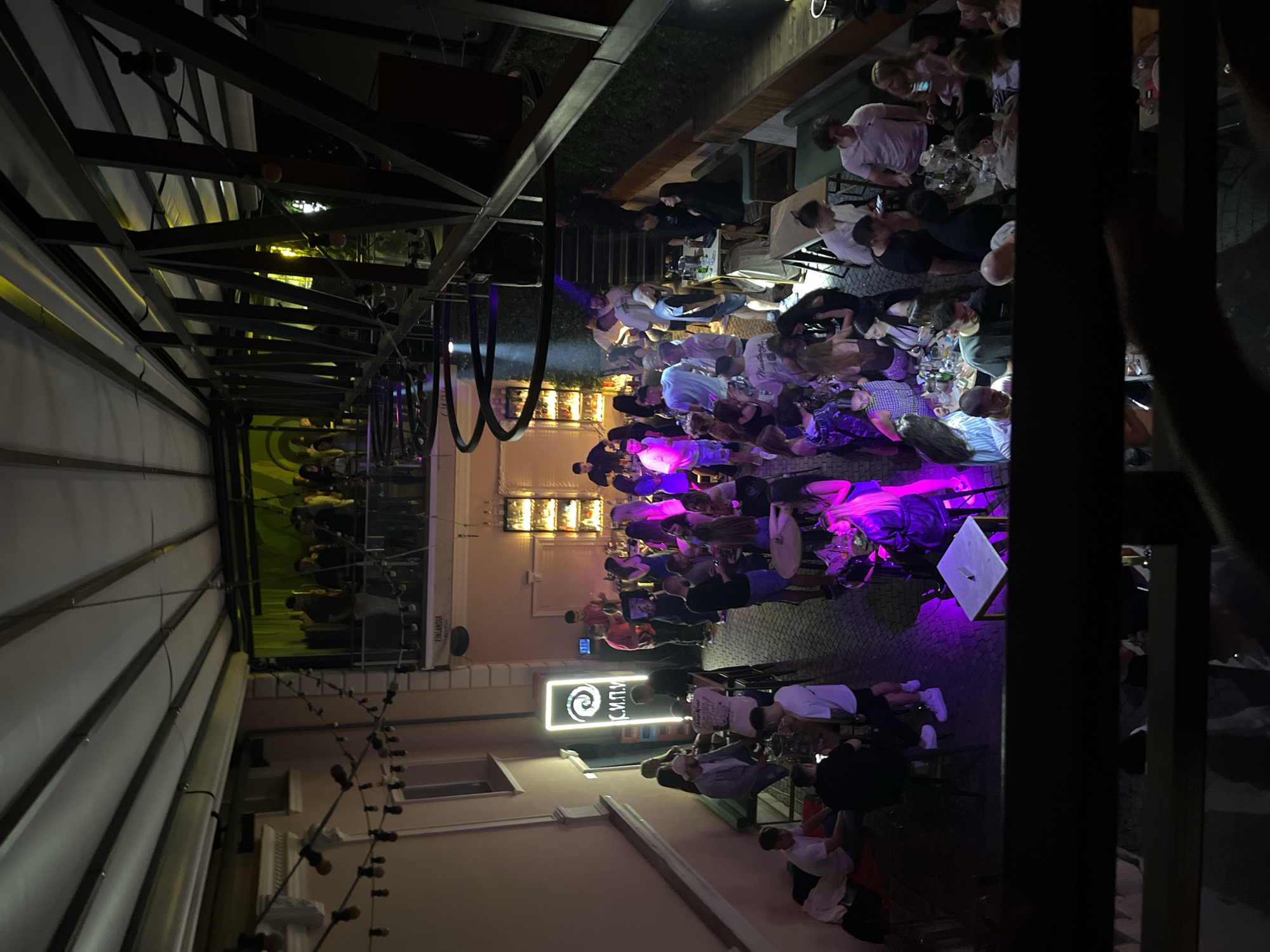 The image depicts a crowded outdoor rooftop bar or club scene at night, likely in Plovdiv, Bulgaria, as suggested by the neon sign "С.И.П.У." which is Cyrillic. The venue is multi-level, with people gathered on the ground floor and a second-level balcony area. String lights hang overhead, and various neon lights and spotlights illuminate the space, casting purple and yellow hues.
On the ground level, there are numerous tables and seating areas, with people standing and seated, socializing, drinking, and some appearing to be smoking shisha. A bar area is visible with bottles and glasses. On the upper level, a group of people are gathered around what appears to be a stage or performance area with yellow lighting.
The architectural style includes brickwork on the walls, metal structures supporting the roof, and a covered ceiling. The overall atmosphere is lively and social, characteristic of a nightlife establishment.