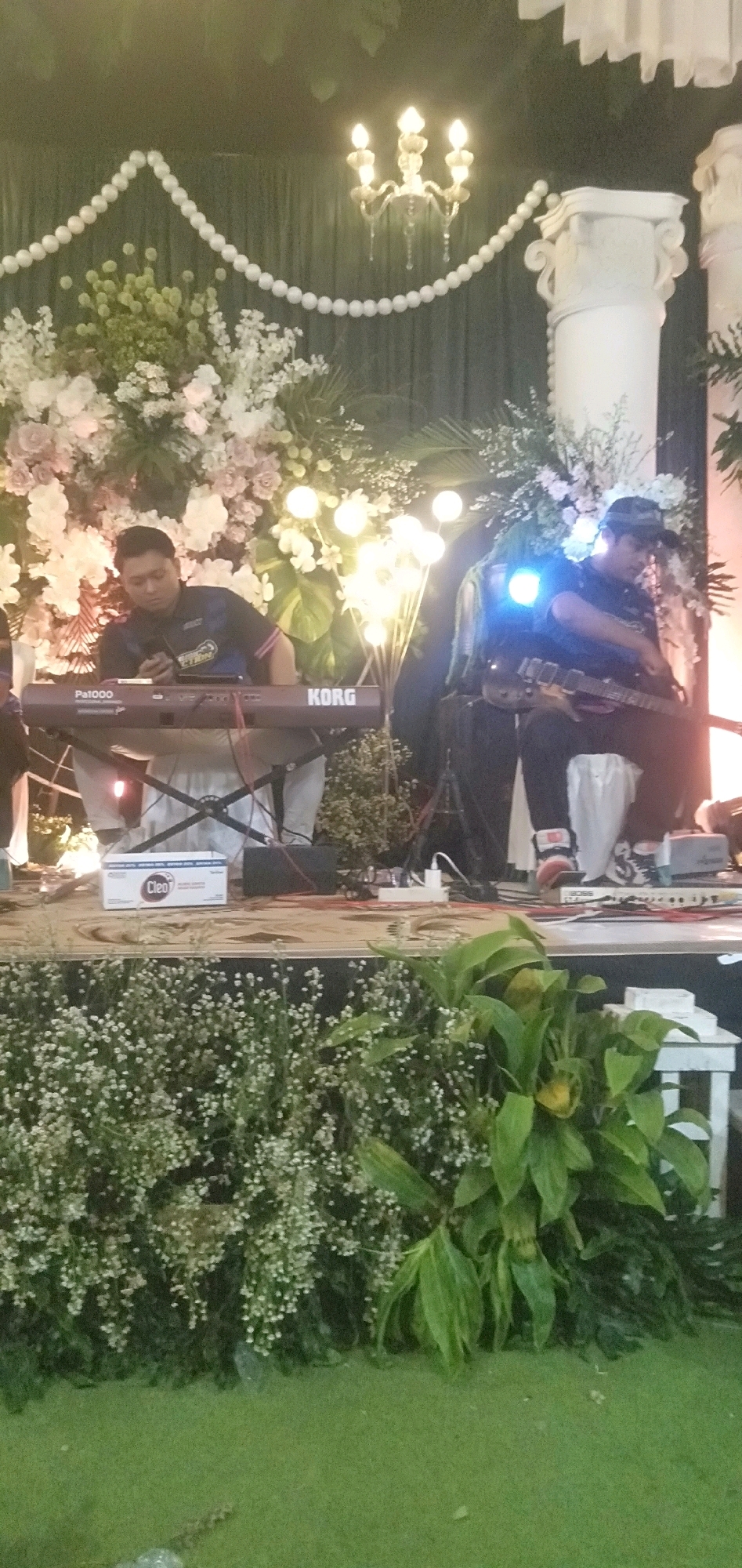 The image depicts an outdoor event, likely a wedding reception or performance, at a venue in Plemahan, Indonesia. A stage is set up with a dark green curtain backdrop, adorned with white pearl-like strings and ornate white columns on either side. The stage is further decorated with abundant floral arrangements featuring white and pink roses, as well as smaller white flowers and greenery. Soft, warm lighting emanates from decorative chandeliers and globe-shaped lights strategically placed among the flowers.
In the foreground, the stage is covered with a patterned carpet. Two musicians are present. On the left, a man is seated and playing a digital keyboard, a KORG Pa1000. He is wearing a dark t-shirt with a colorful logo and light-colored pants. In front of the keyboard, there is a white box with the "Cleo" logo.
On the right side of the stage, another man is standing and playing an electric guitar. He is wearing a dark t-shirt and a dark baseball cap, and dark pants. He is illuminated by a bright blue light, suggesting stage lighting. His guitar is dark-colored.
The stage is bordered by lush green foliage and small white flowers in the foreground, which transitions to a bright green, carpet-like ground cover in the very bottom of the image. The overall atmosphere suggests a celebratory and well-decorated occasion.