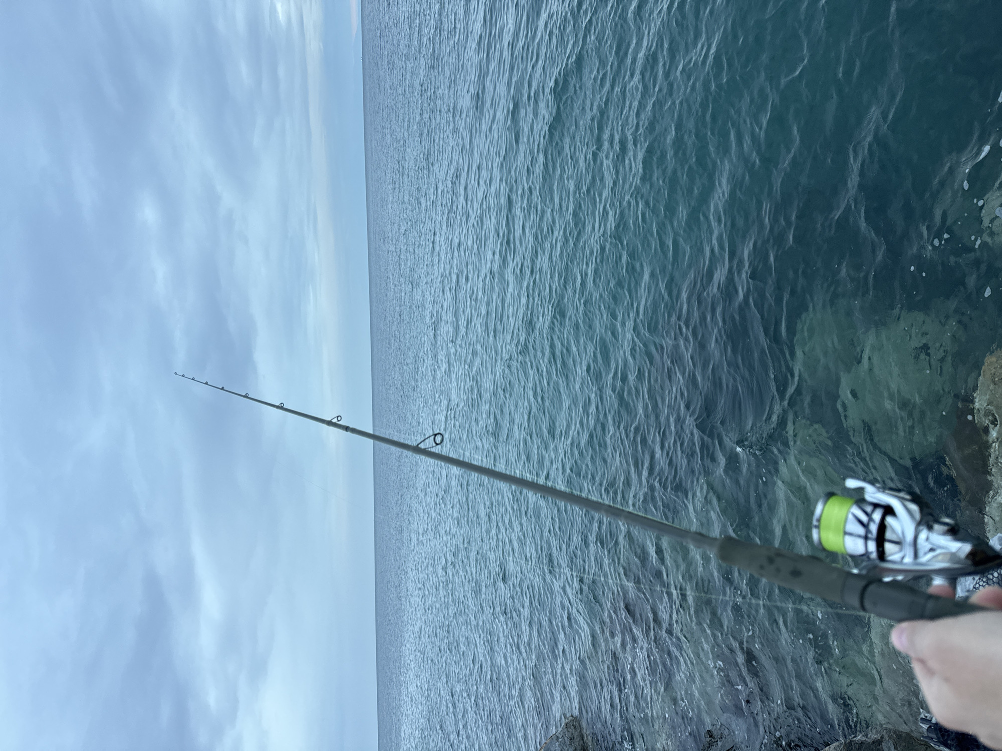 A person is holding a fishing rod on a rocky outcrop, presumably in Νέα Καλλικράτεια, Greece. The ocean stretches out to the horizon under a cloudy sky. The fishing rod is pointed towards the water, and the reel is visible near the handle.