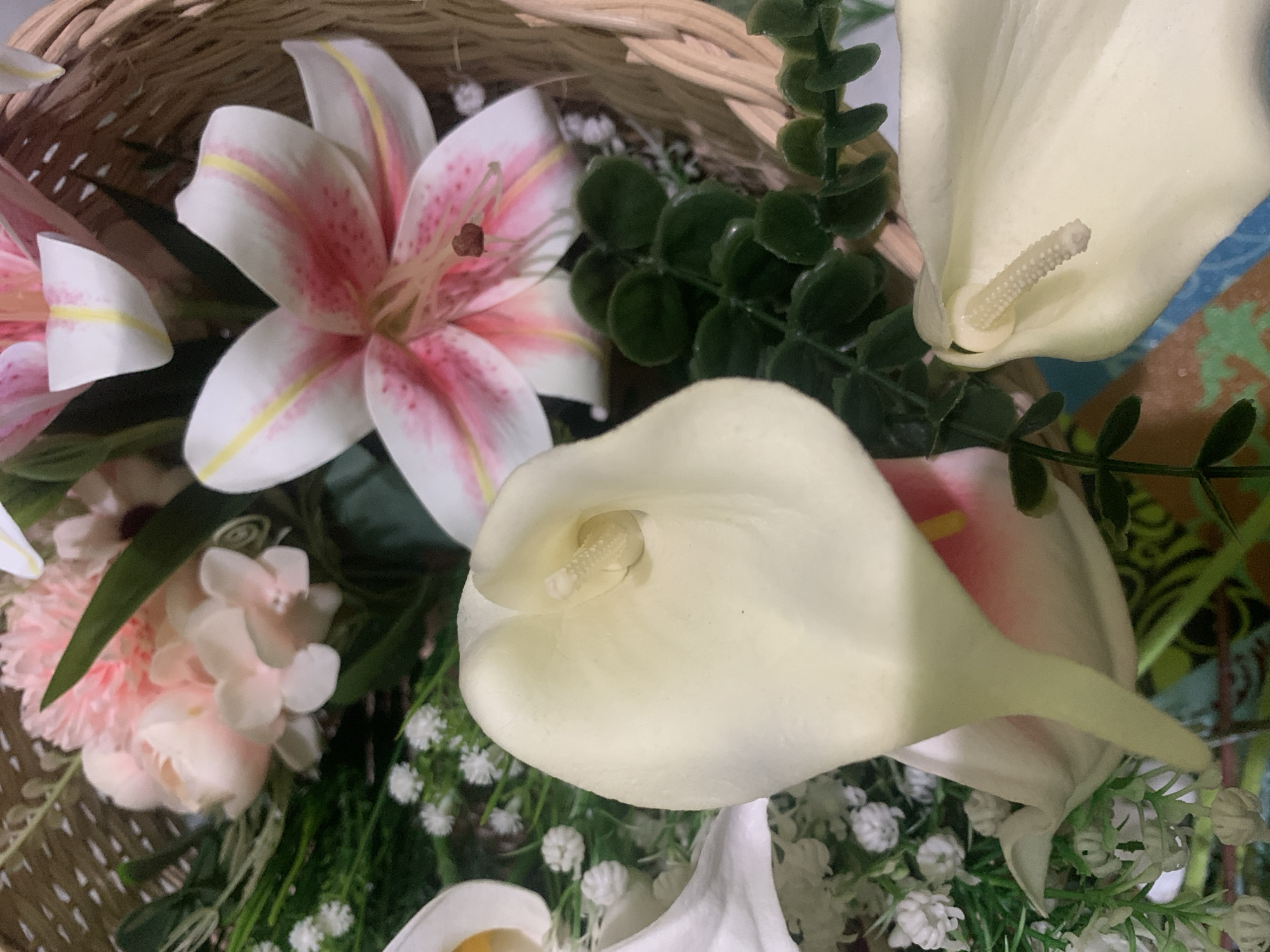 The image displays a close-up view of a floral arrangement in a woven basket. The arrangement features several types of artificial flowers, including large white calla lilies with prominent spadices, pink and white lilies with speckled petals, and smaller pink and white blossoms. Green foliage and tiny white filler flowers are interspersed throughout. The background is blurred, showing hints of a basket weave and a colorful patterned surface. The lighting is soft, creating a natural and gentle illumination of the flowers. There are no people or animals present in the image, and no specific landmarks or environmental features are visible that would indicate a location in Kuala Pembuang, Indonesia.