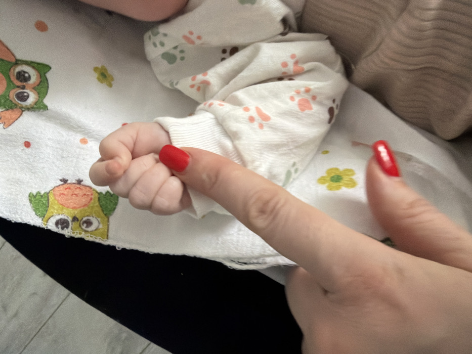A baby's hand, clad in a white patterned sleeve, is gently touched by a woman's index finger adorned with red nail polish. The baby's hand is small and clenched, while the woman's finger is extended towards it. The background features a patterned white fabric with owl and floral designs, and a glimpse of black fabric at the lower edge. The overall scene is intimate and tender.
