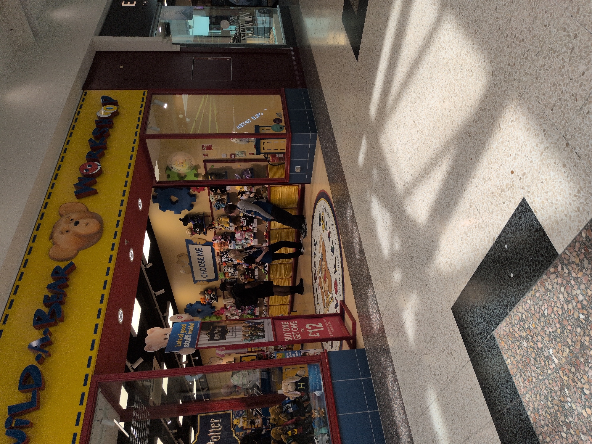 The image shows the exterior of a "Build-A-Bear Workshop" store located within a shopping center. The store's facade is predominantly yellow with red accents and features large glass windows displaying stuffed animals and merchandise. The store's name, "BUILD-A-BEAR WORKSHOP," is prominently displayed in red and blue lettering across the top of the building. Inside the store, several people are visible, including employees and customers interacting with the displays.

A promotional sign outside the store reads "BUY ONE GET ONE FOR £12". The floor of the shopping center is made of speckled tile, with shadows from overhead lighting casting patterns across the ground. The overall environment appears to be a brightly lit indoor shopping mall.

This particular Build-A-Bear Workshop is located in the Telford Shopping Centre in Telford, United Kingdom. The shopping centre is a major retail destination in the region.