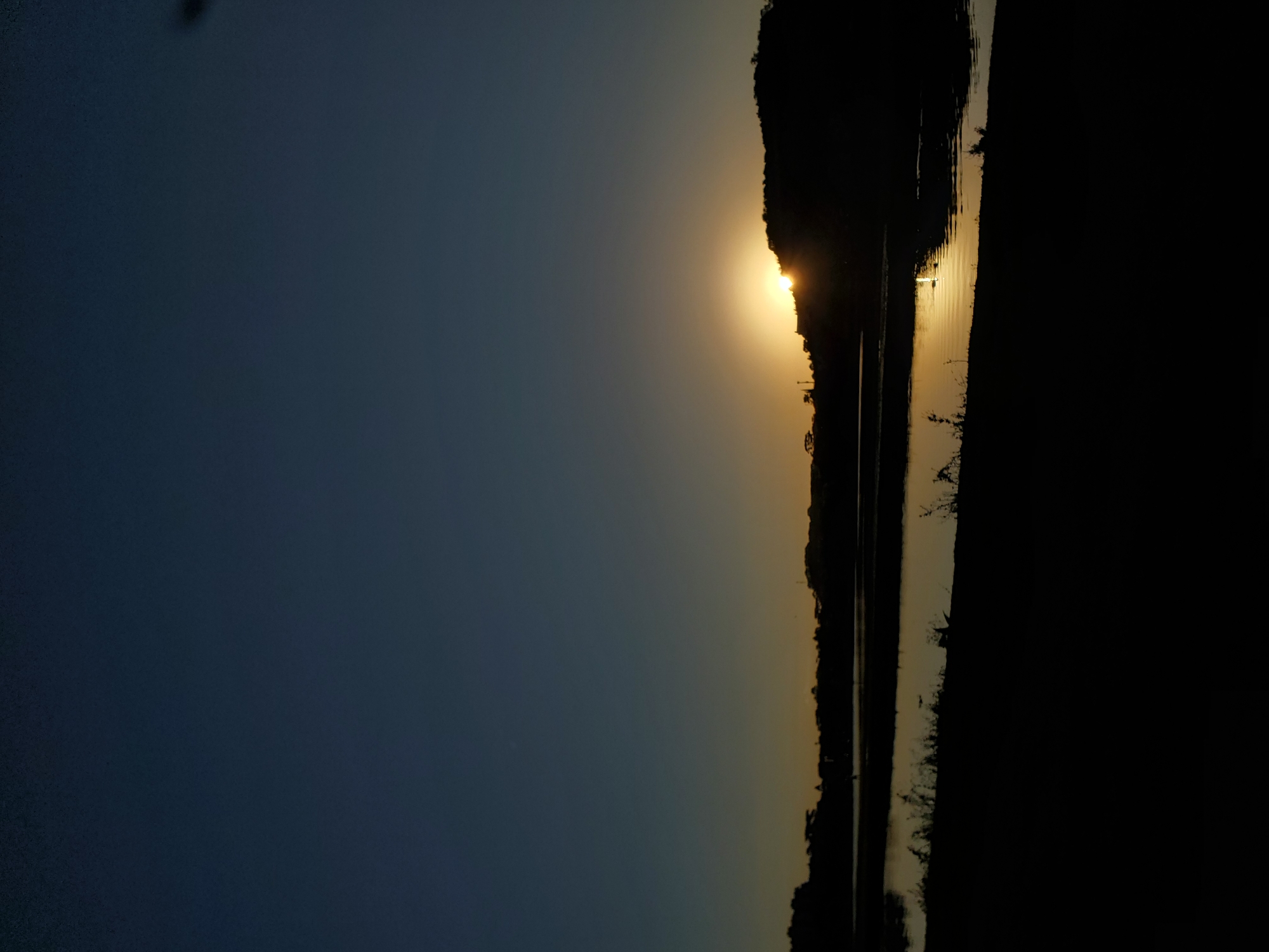 The image depicts a sunset over a body of water, likely a lake or river, in Curitiba, Brazil. The sky is a deep blue that transitions to a pale orange near the horizon. Silhouetted against the setting sun, which is partially obscured by a dark, hilly landscape, are trees and what appear to be structures. The water reflects the warm colors of the sunset, creating shimmering golden hues on its surface. In the foreground, the land is also silhouetted in darkness, with some vegetation visible along the water's edge. No people or animals are clearly discernible. The overall atmosphere is tranquil and serene, characteristic of a peaceful evening in an urban or suburban natural setting.