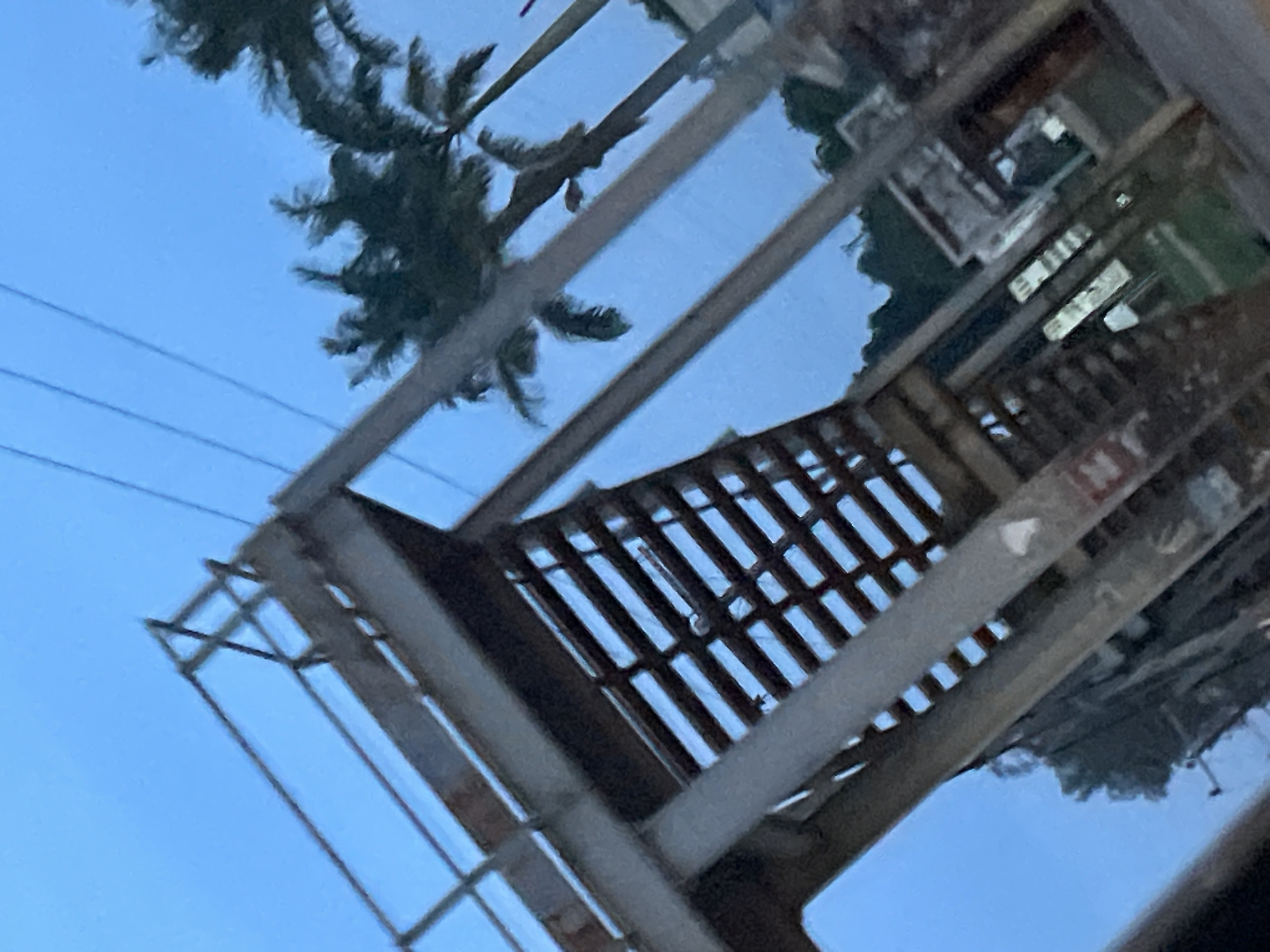 The image shows an outdoor scene featuring an elevated metal walkway or bridge with visible steps and railings. Palm trees are visible in the background, suggesting a tropical or subtropical environment. Overhead power lines are also present, indicating an urban or semi-urban setting. The lighting suggests it is either dawn or dusk. There are no people or identifiable actions depicted. The scene appears to be located in the city of Jos, Nigeria, based on the provided context.