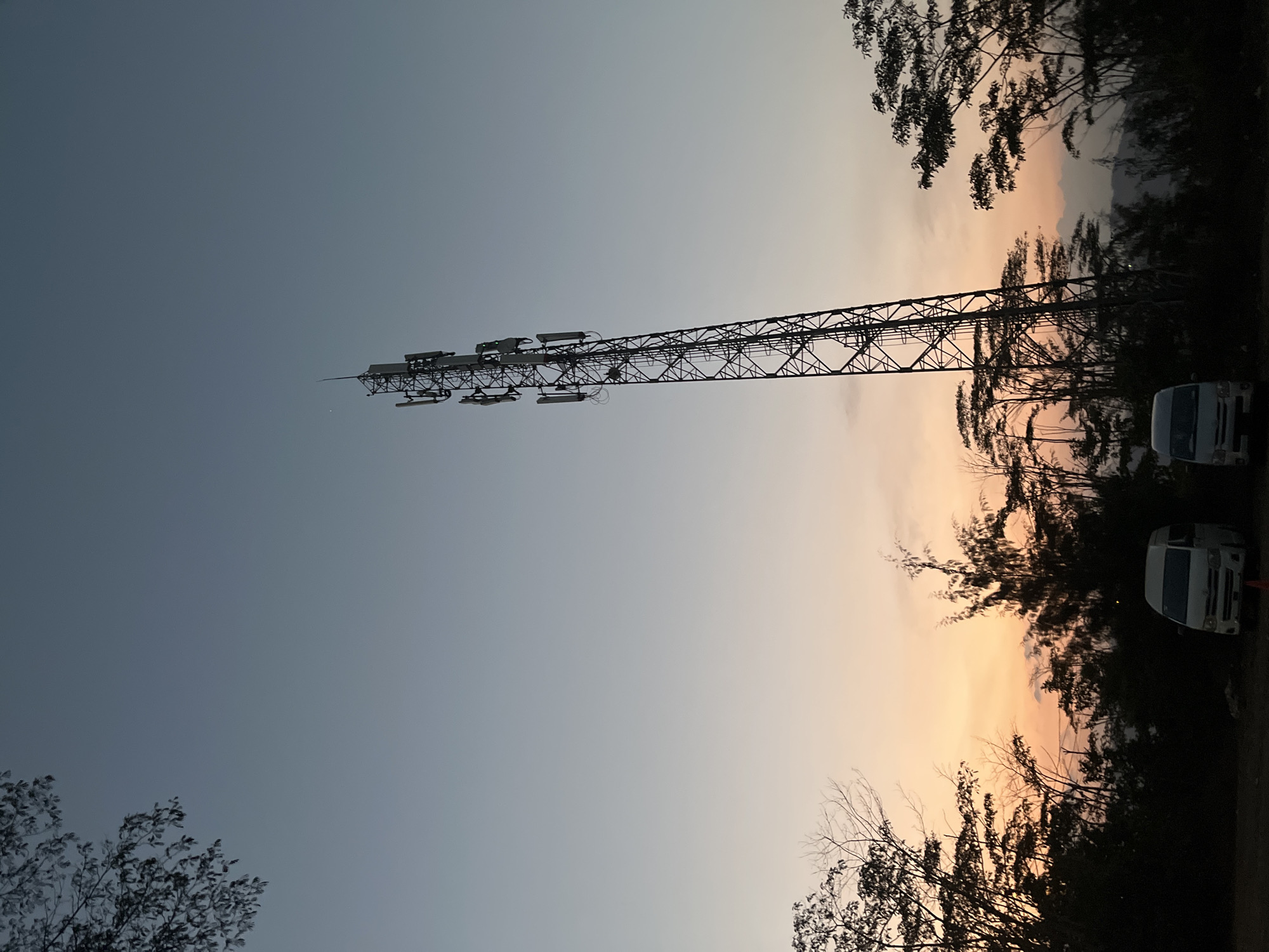 The image shows a cell tower silhouetted against a twilight sky. The sky transitions from a pale blue at the top to a warm orange and pink hue near the horizon, indicating either sunrise or sunset. Several trees with dark foliage are visible in the foreground and middle ground, with some bare branches intermingled. Two white vans are parked in the lower left portion of the image. The overall scene is set outdoors, likely in a rural or semi-rural area, given the presence of trees and the open sky. No people are clearly discernible in the image. The location is specified as Lalampu, Indonesia.