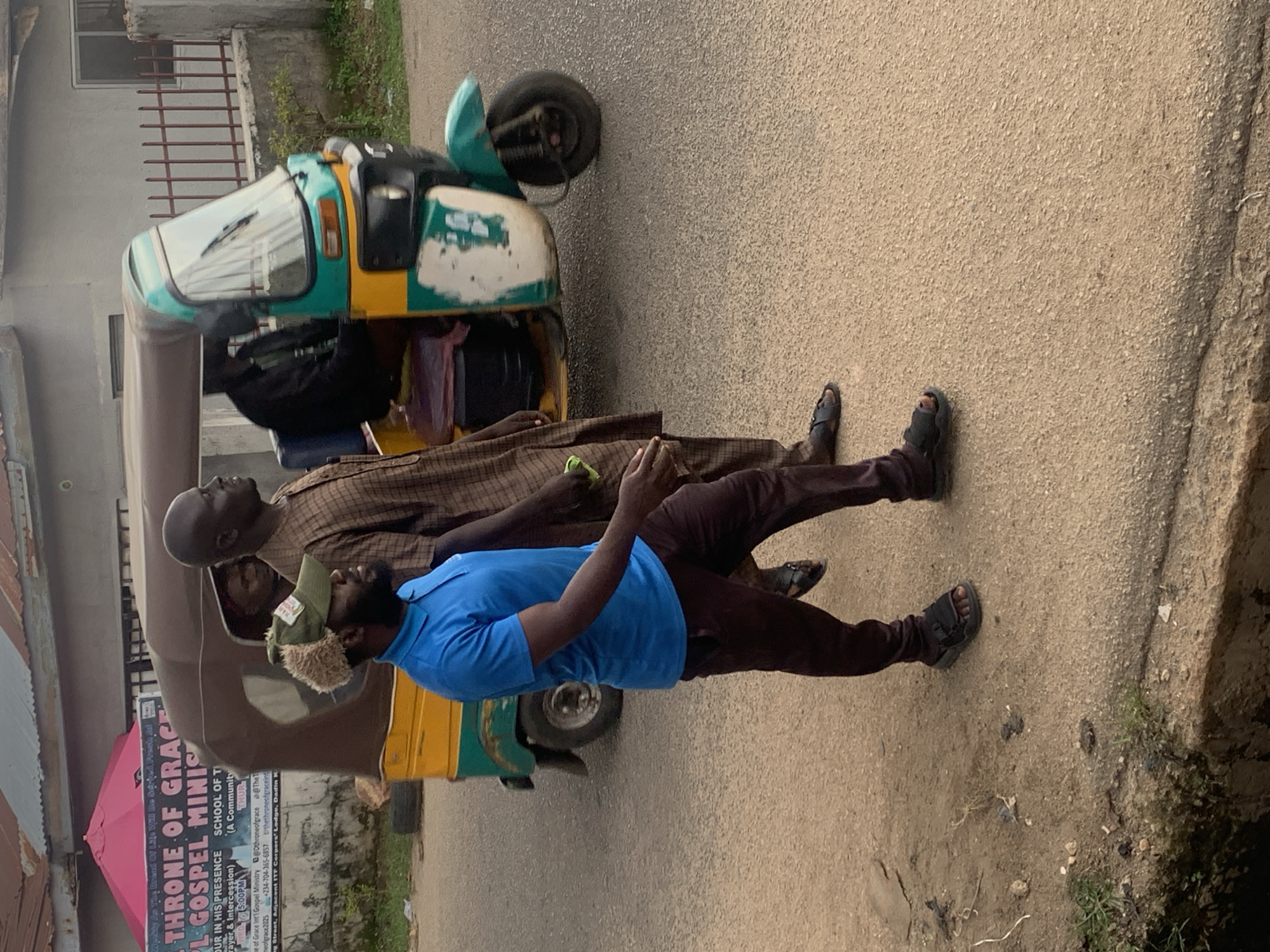 The image shows two men standing on a paved surface, likely a road, with a three-wheeled vehicle, also known as an auto-rickshaw or tuk-tuk, parked nearby. One man is wearing a brown checkered outfit and the other is in a blue t-shirt and dark pants. The man in blue is wearing a distinctive cap with fuzzy ear flaps. Both men are looking upwards, with seemingly neutral expressions.
The auto-rickshaw is brightly colored, with green and yellow sections, and appears well-used. A pink umbrella is visible leaning against the side of the vehicle, partially obscuring some text. The text, partially visible on the side of the auto-rickshaw, includes phrases like "THRONE OF GRACE" and "GOSPEL MINIS," suggesting it might be associated with a religious organization. Other visible text includes "SCHOOL OF T," "OUR IN HIS PRESENCE," and "Payer & Intercession." Contact information and a street name are also present.
The setting appears to be an urban or semi-urban environment in Jos, Nigeria, with a building visible in the background. The lighting suggests it is daytime, possibly overcast given the lack of strong shadows. The ground is paved with asphalt, and there are patches of grass and debris along the side. The overall scene depicts a moment of pause or interaction on a street, potentially related to the religious outreach suggested by the text on the vehicle.