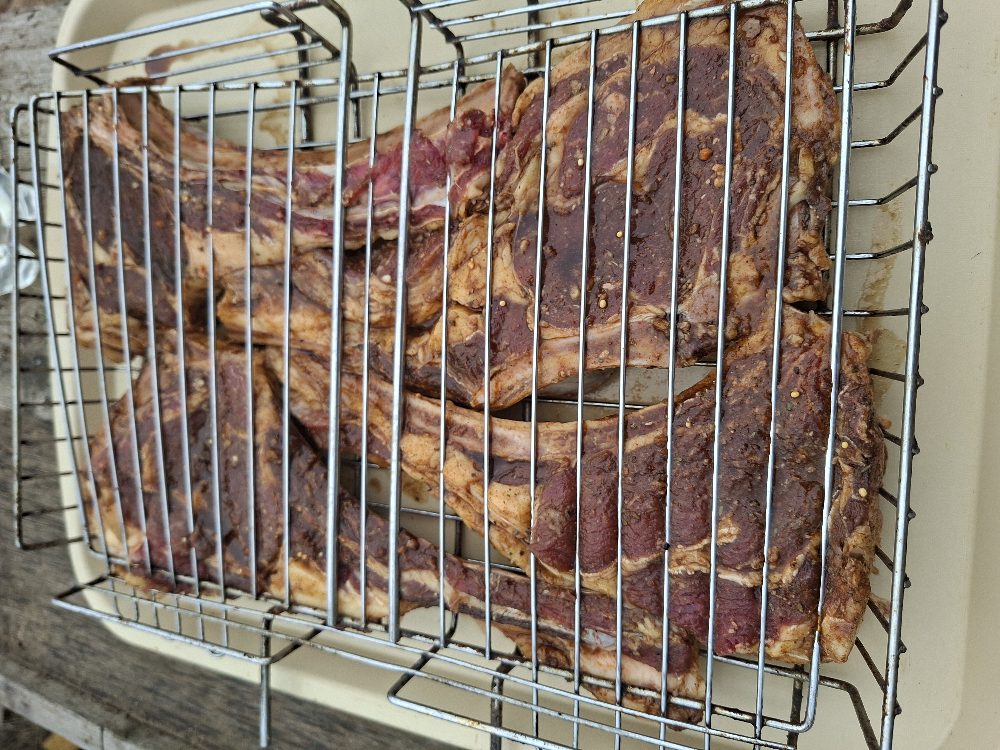 The image shows raw, marinated beef short ribs in a wire grilling basket. The basket sits on a beige tray, possibly to contain drips. The ribs are cut thinly across the bone and heavily coated in a dark marinade with visible seasonings. The background consists of weathered wooden surfaces, suggesting an outdoor setting. The location is not identifiable.