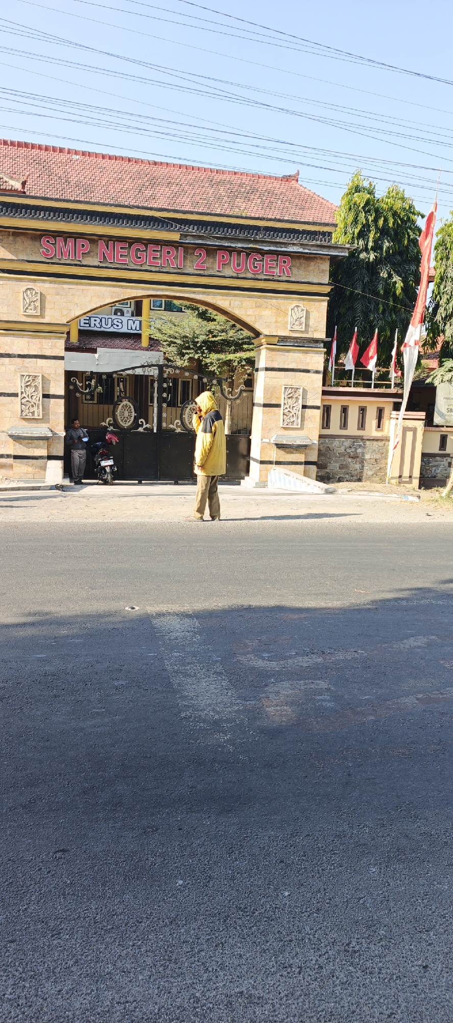 The image shows the entrance gate to SMP Negeri 2 Puger, a school in Kasiyan Timur, Indonesia. A person wearing a yellow hooded jacket and brown pants is standing in front of the gate, appearing to be talking on a cell phone. To the left, another person in a gray shirt and pants stands near a motorcycle. The gate itself is made of wrought iron with decorative elements, and above it, red lettering reads "SMP NEGERI 2 PUGER". The building behind the gate has a red tile roof and is decorated with stone-like materials. Several Indonesian flags are visible to the right of the gate. The scene is set on a street in front of the school, with an asphalt road and clear sky overhead.