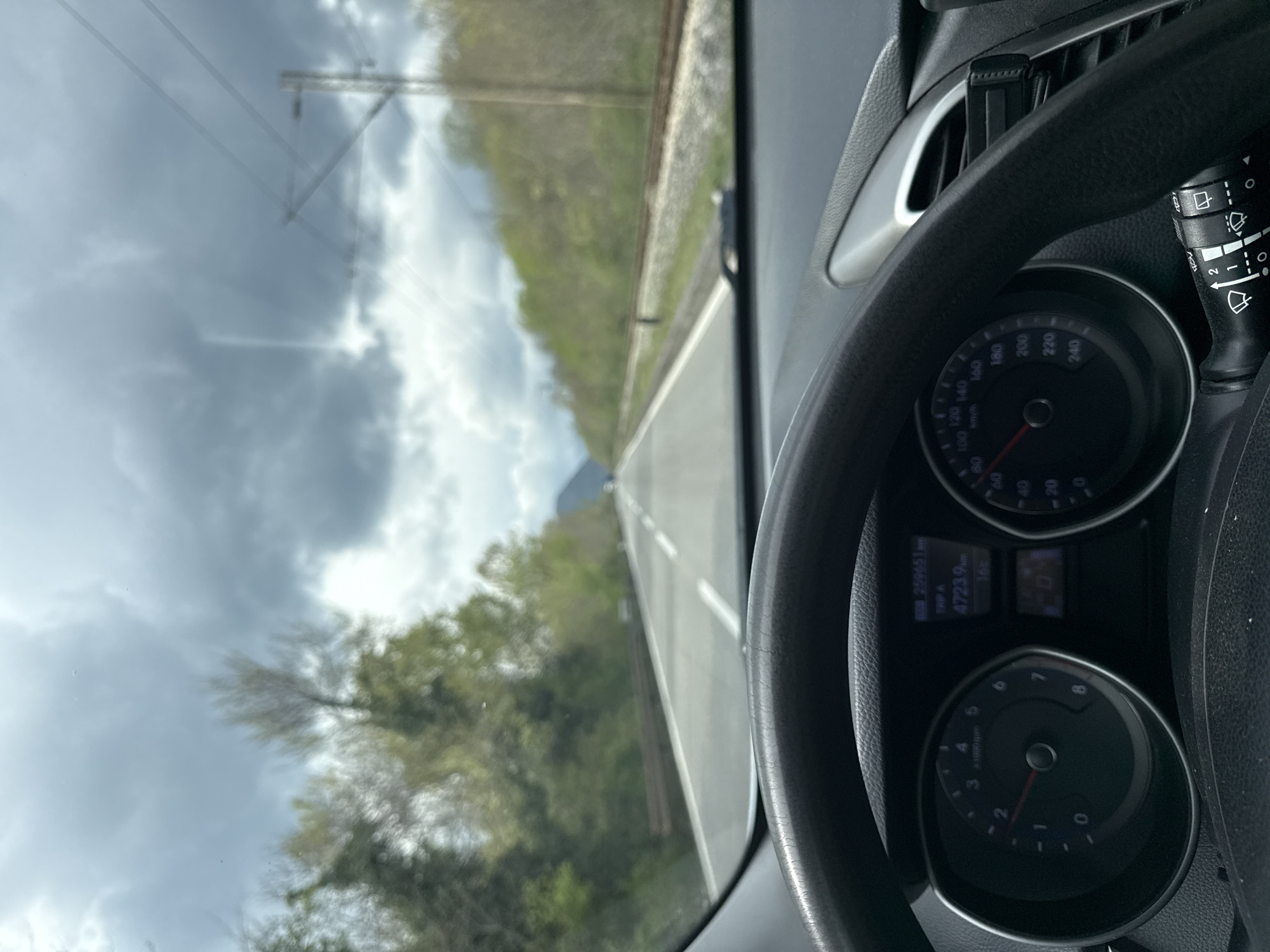 The image depicts a first-person perspective from within a moving vehicle, located in the city of Bistrice, Montenegro. The lower section of the frame is occupied by a black, textured steering wheel. Behind it, the vehicle's instrument cluster displays a tachometer on the left, with its needle indicating approximately 1100 RPM, and a speedometer on the right, showing a speed of approximately 90 km/h. A central digital display reveals an odometer reading of 259651 km, a trip meter indicating 4723.9 km, and an ambient temperature of 16°C. A black control stalk for windshield wipers is visible to the right of the steering wheel, next to a partially visible silver-trimmed air vent that contains a black phone holder.
Through the vehicle's windshield, a two-lane asphalt road is visible, extending into the distance under an overcast sky filled with dark grey clouds. The road features a solid white line separating opposing directions of traffic and dashed white lines marking individual lanes. To the left of the road, a metal guardrail runs parallel, backed by a dense array of green and light-green deciduous trees and foliage. To the right of the road, a single set of railway tracks is present on a gravel bed, also flanked by similar deciduous trees and foliage. A utility pole, likely supporting overhead lines, is discernible further down the railway. Distant hills or mountains are faintly visible on the horizon, partially obscured by the cloud cover. The vehicle is in motion, indicated by the speedometer and tachometer readings. No individuals are explicitly visible.