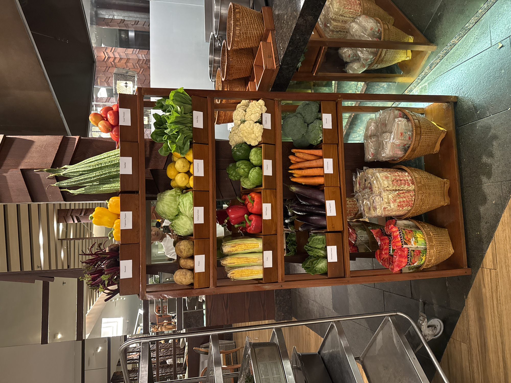 The image shows a tiered wooden display rack filled with fresh vegetables. The rack is positioned in what appears to be a market or a restaurant buffet, given the presence of stainless steel carts and kitchenware in the background. On the top shelf, there are beets, yellow bell peppers, and tomatoes. Below that, potatoes, iceberg lettuce, lemons, and bok choy are displayed. The next shelf features corn, bell peppers, chayote, and cauliflower. The fourth shelf contains romaine lettuce, long eggplant, carrots, and broccoli. The bottom shelf holds packaged noodles or snacks in wicker baskets. The overall environment suggests a well-organized and abundant selection of produce. The location is identified as Kuta Selatan, Indonesia, hinting at a tropical setting.