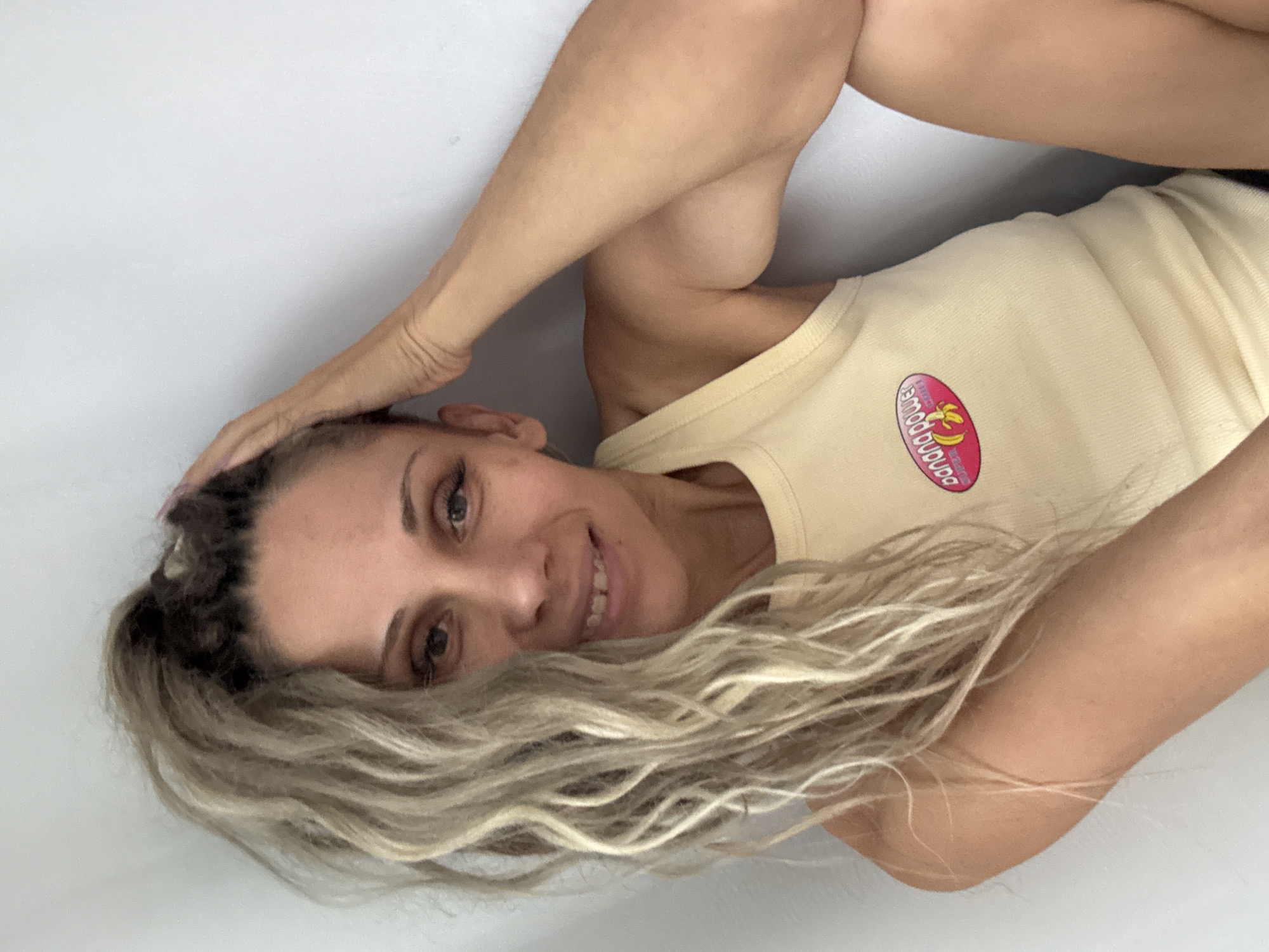 In Rusanovo, Russia, a woman with blonde, wavy hair pulled into a ponytail, is sitting with her arm raised to her head. She is wearing a light yellow tank top with a pink oval logo containing the text "BANANA POWER" and a banana graphic. She is looking towards the camera with a slight smile. The background is a plain, light-colored wall.