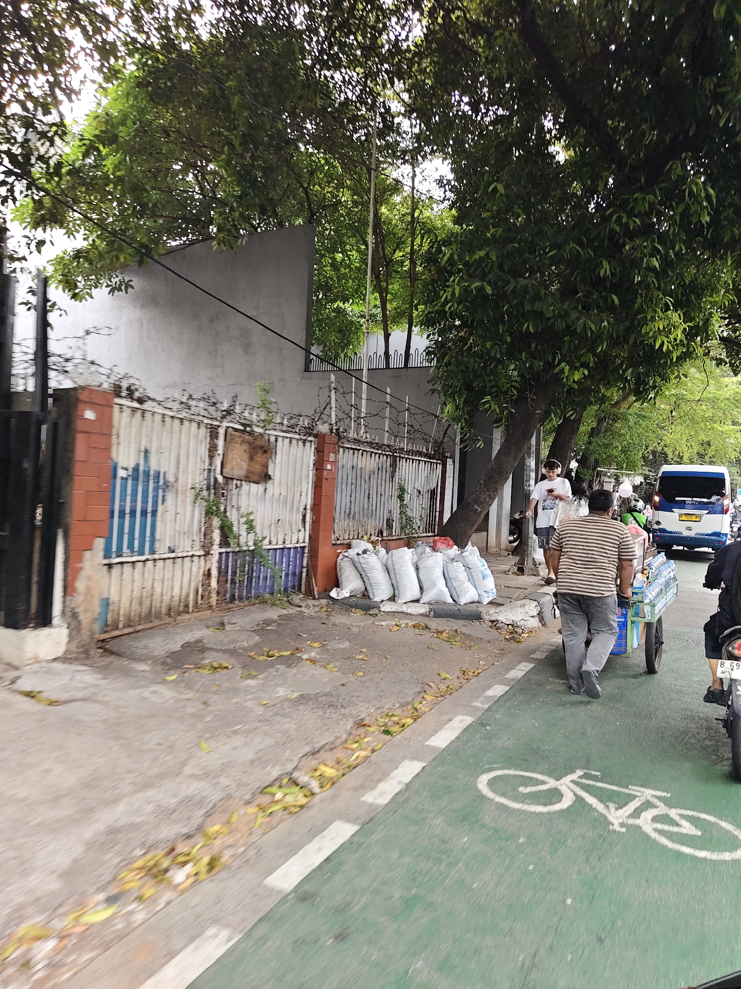 A slightly blurred, medium-shot street-level photograph, taken from a slightly elevated perspective, captures a scene on Jalan Kyai Haji Hasyim Ashari in West Jakarta, Indonesia. The image depicts a busy roadside with a distinct bike lane, marked by a green surface and a white bicycle symbol. On the right side of the frame, a man in a striped shirt and gray pants pushes a cart laden with bottled water, moving along the bike lane. Behind him, a motorcycle rider is partially visible. To the left of the bike lane, a sidewalk runs alongside a weathered metal fence topped with barbed wire, which encloses a property with a concrete wall. Several white sacks are stacked neatly on the sidewalk near the fence. Further left, trees with dense green foliage overhang the scene, creating a dappled light effect. In the background, a white van with blue accents and an Indonesian license plate is parked on the road. The overall impression is one of a typical urban street environment in Jakarta.