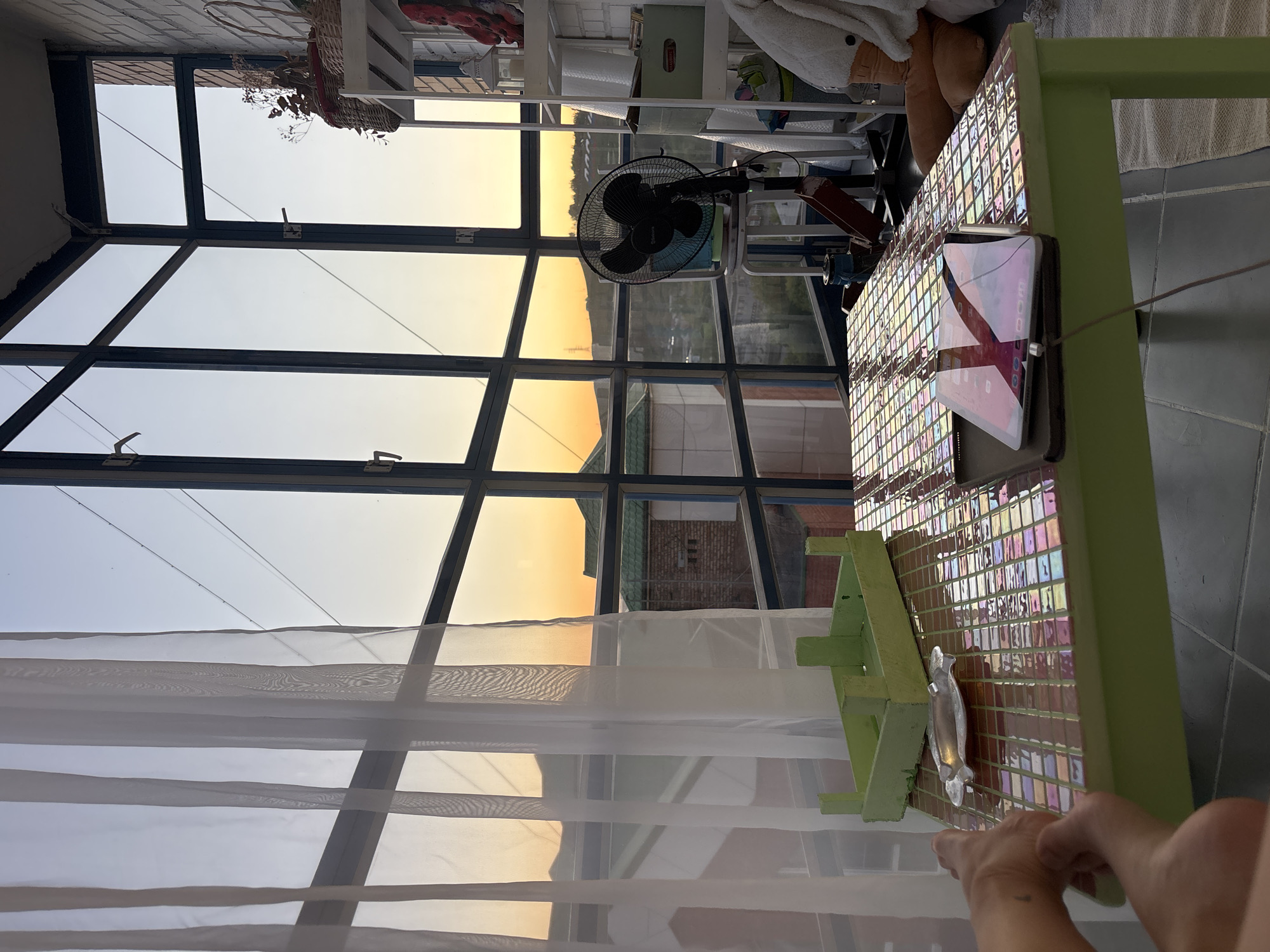 This media file captures an indoor scene, likely a sunroom or enclosed balcony, at either sunrise or sunset, with a prominent view through large grid-paned windows. The sky outside is illuminated with a warm, golden glow, suggesting clear weather.
In the foreground to the right, a person's hands are visible, resting on or near a table. The table features a mosaic top composed of colorful, square tiles and has a light green frame. A tablet computer rests on the table, its screen reflecting ambient light and displaying various icons; a faint reflection that might read "DOOM" or similar text is visible. A small ashtray or dish is also present on the table.
Centrally placed near the windows is a black electric fan, bearing the brand name "Sakura" on its cover. White, sheer curtains partially cover the lower window sections, diffusing the light entering the room. In the upper right background, white shelving units hold various items, including a woven basket filled with dried plants or flowers, and a folded white towel or blanket.
The view through the windows extends to a horizon with distant buildings, silhouetted against the golden sky. The scene evokes a sense of calm and tranquility. The provided location context for this image is Krasnoyarsk, Russia.