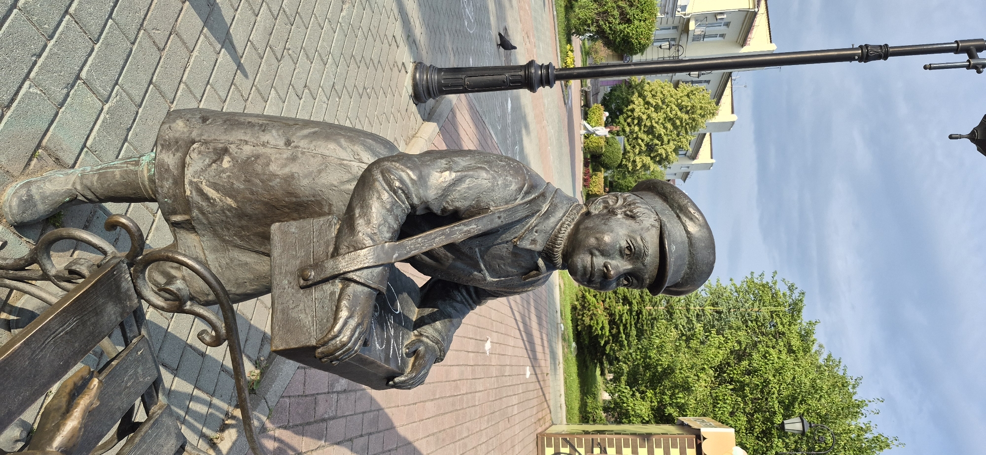 A bronze statue of a young boy wearing a cap, coat, and satchel is positioned on a paved walkway in the city of Ussuriysk, Russia. The boy is seated and holding a rectangular box. A lamppost is to the left of the statue, and a park bench is to the right. In the background, there are buildings, trees, and a few people walking in the distance. The sky is cloudy. The ground is paved with bricks, and there is a pigeon on the ground near the lamppost.