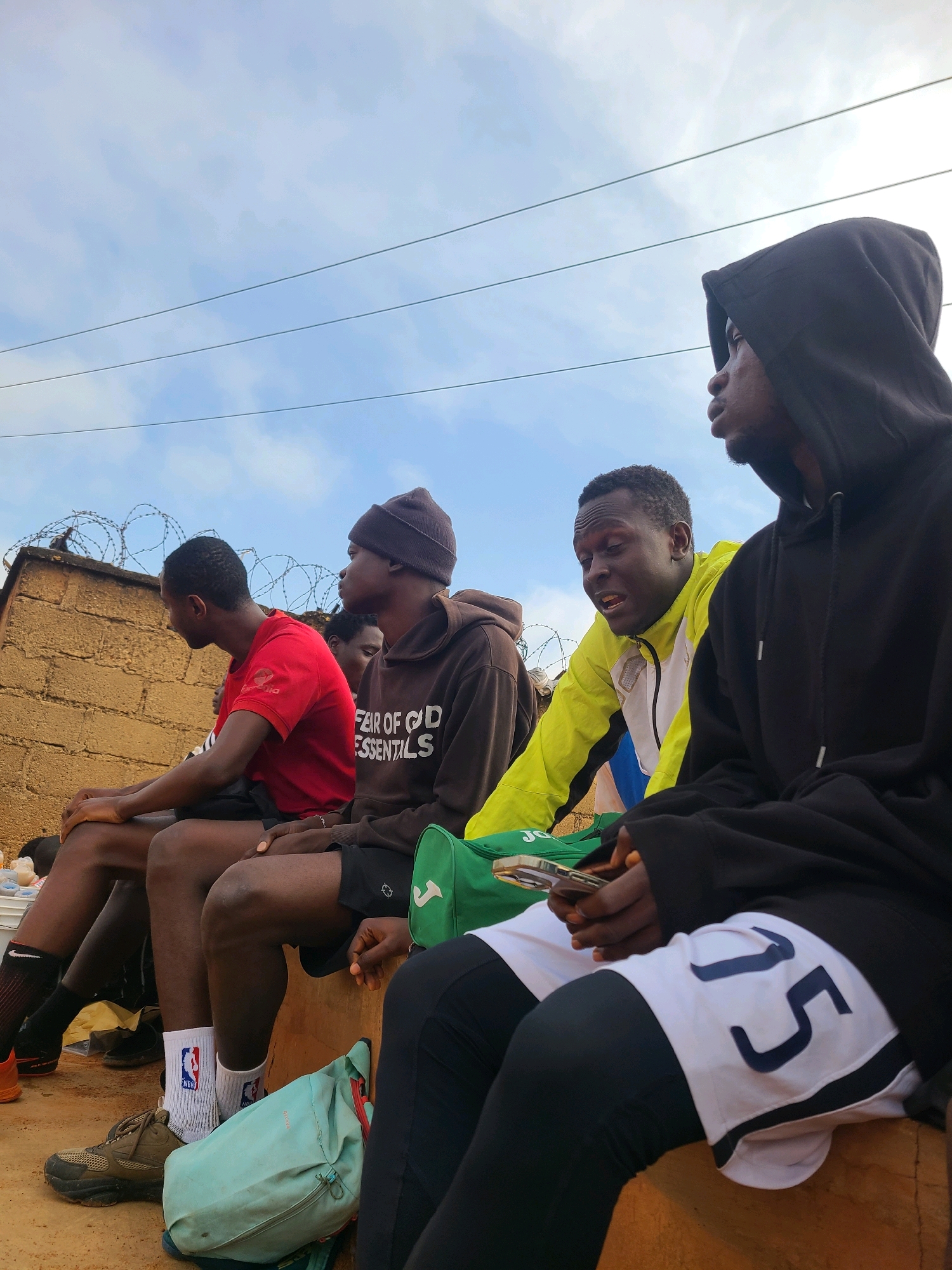 The image depicts a group of young men sitting outdoors, possibly in a rural or semi-urban setting in Barakin Lerawi, Nigeria. The sky is cloudy, suggesting it could be overcast or late afternoon. The men are dressed casually, some in athletic wear. One man in the foreground is wearing a black hooded sweatshirt and white shorts with the number "75" in dark blue. He is holding a smartphone. Next to him, a man in a bright yellow and white jacket is speaking, appearing animated. To his left, a man in a red t-shirt is looking away. Another man wearing a brown beanie and a brown hoodie with the text "FEAR OF GOD ESSENTIALS" is also present. A light blue duffel bag and a green sports bag are visible on the ground near the men. The background includes a brick wall topped with barbed wire, and power lines are visible against the sky. The overall impression is of a casual gathering, possibly a break during some activity or simply a moment of repose. The expressions and postures suggest a relaxed atmosphere, with the man in the yellow jacket appearing to be engaged in conversation.