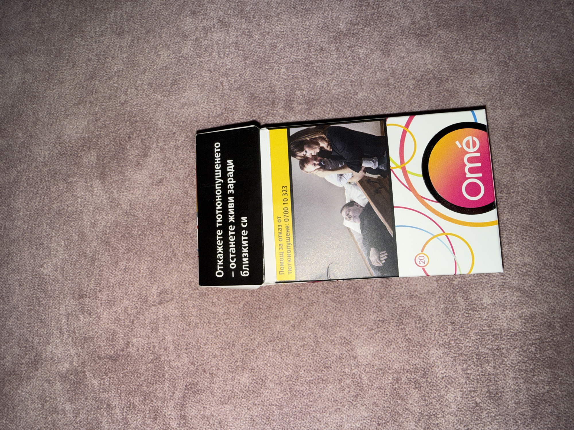 A pack of Omé brand cigarettes is positioned centrally on a textured, light brown surface, likely upholstery or carpet, in the city of Unknown, Unknown. The cigarette pack features a prominent black warning label at the top, stating in white Bulgarian text: "Откажете тютюнопушенето – останете живи заради близките си" (Quit smoking - stay alive for your loved ones). Below this, a yellow strip contains further Bulgarian text: "Помощ за отказ от тютюнопушене: 0700 10 323" (Help to quit smoking: 0700 10 323). The main body of the pack displays a graphic image: a woman, positioned on the right, holds an infant with a pacifier, while a man, partially visible on the left, lies in what appears to be a bed, his hands resting on his chest, suggesting illness or death. The bottom section of the pack is white with colorful, overlapping circular and curved line patterns, featuring the "Omé" logo in white within a gradient pink-to-orange oval. A small circular emblem with the number "20" is visible on the upper left corner of this lower section.
